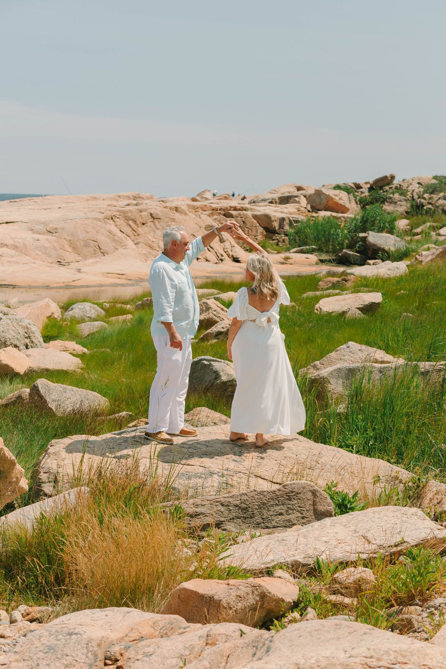 Couple in white outfits standing on rocky coastal landscape with green grass and ocean views on a sunny day.