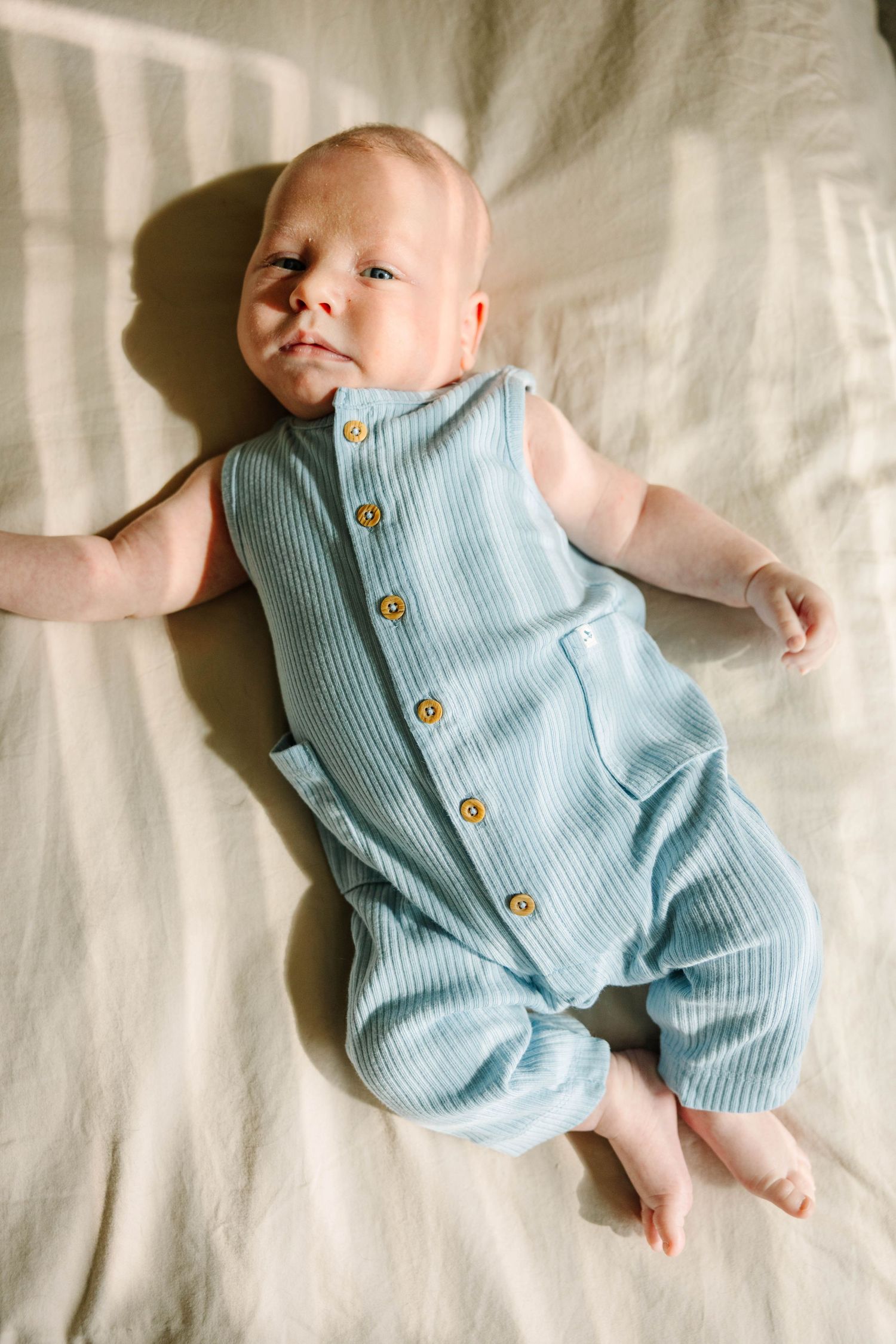 Newborn baby in a light blue ribbed sleeveless romper with yellow buttons, lying on a cream linen surface.