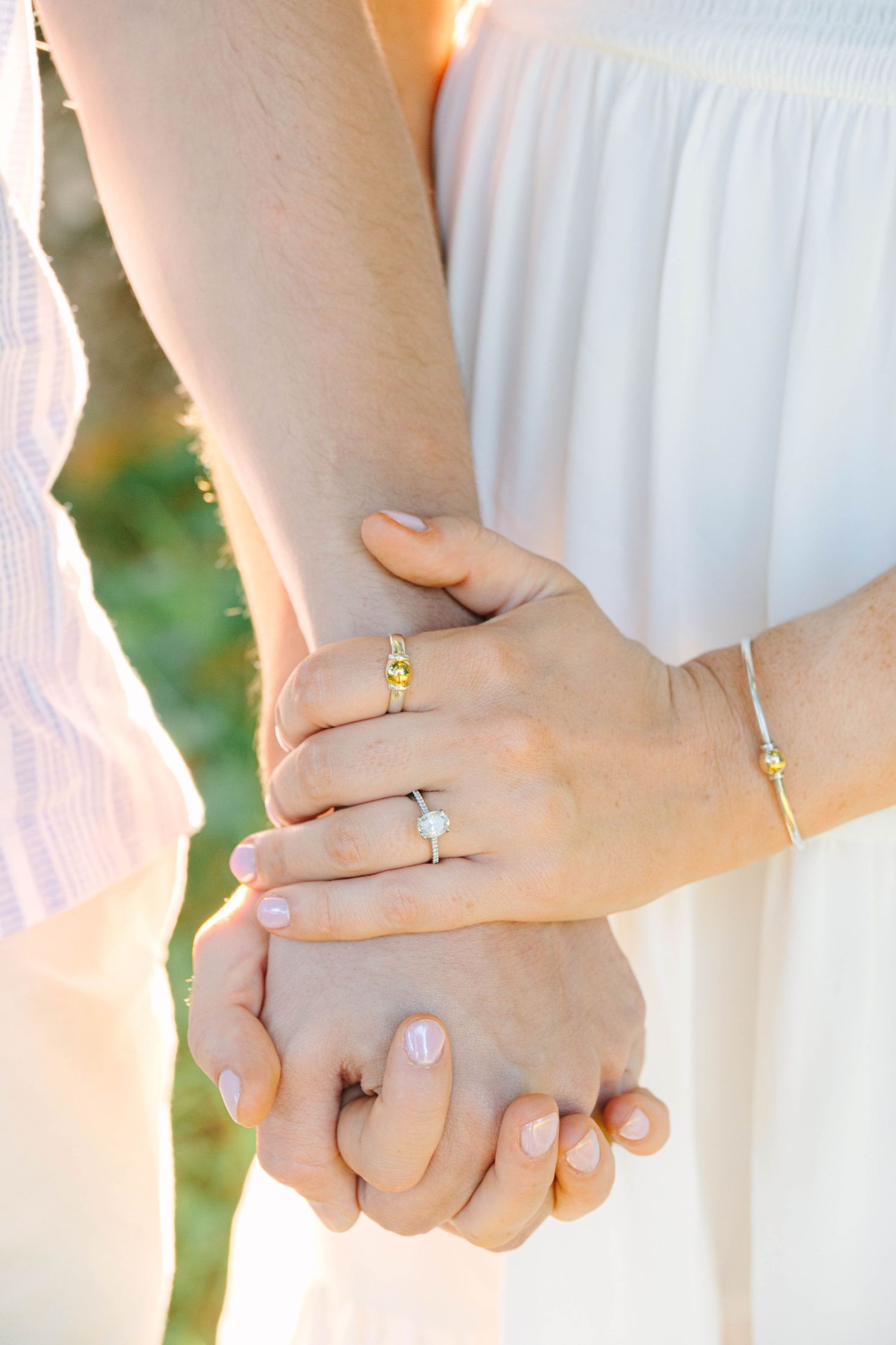 Close-up of couple holding hands, woman wearing diamond engagement ring and gold bracelet in warm sunlight.