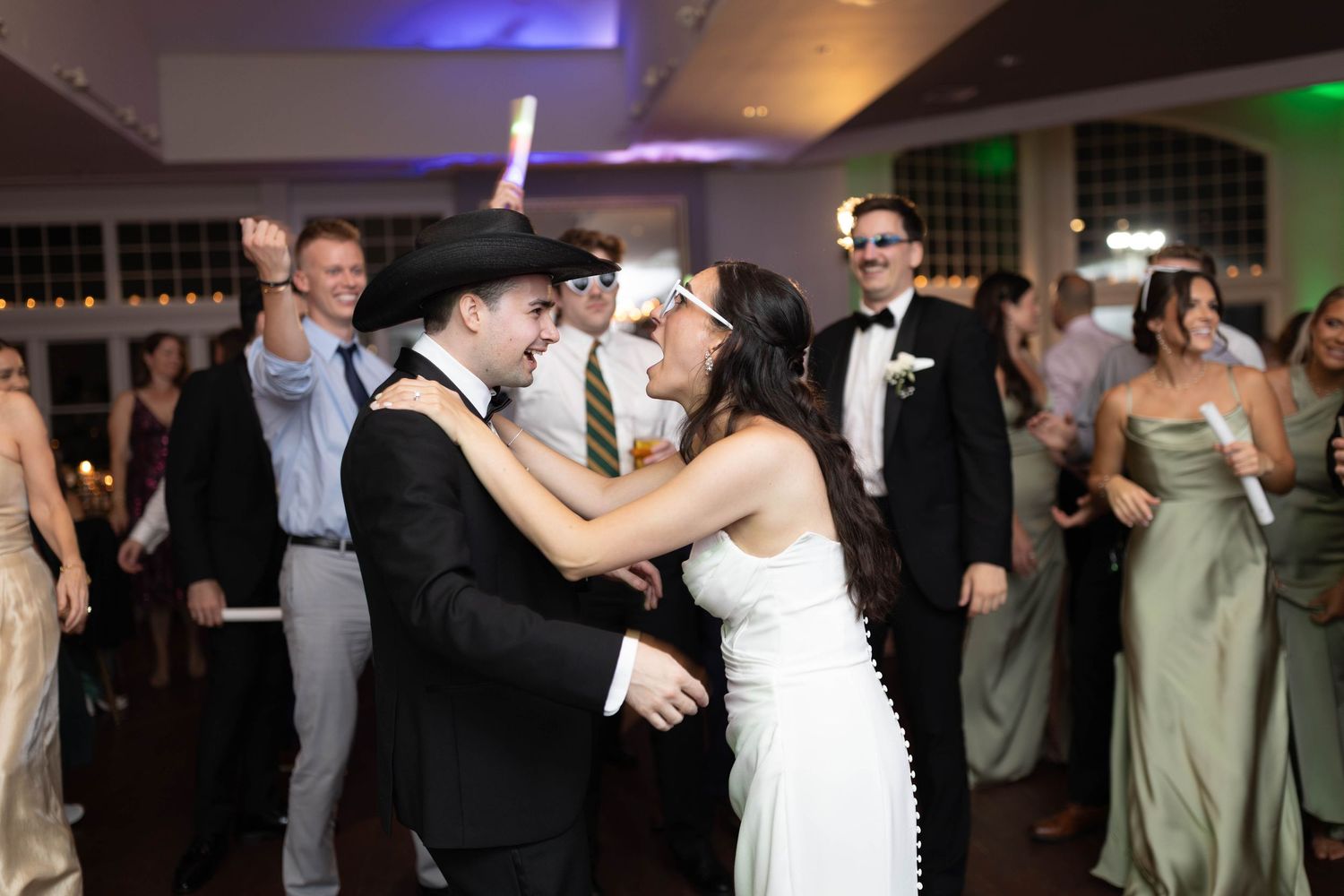 Bride and guests dance joyfully on wedding reception dance floor with colorful uplighting in background.
