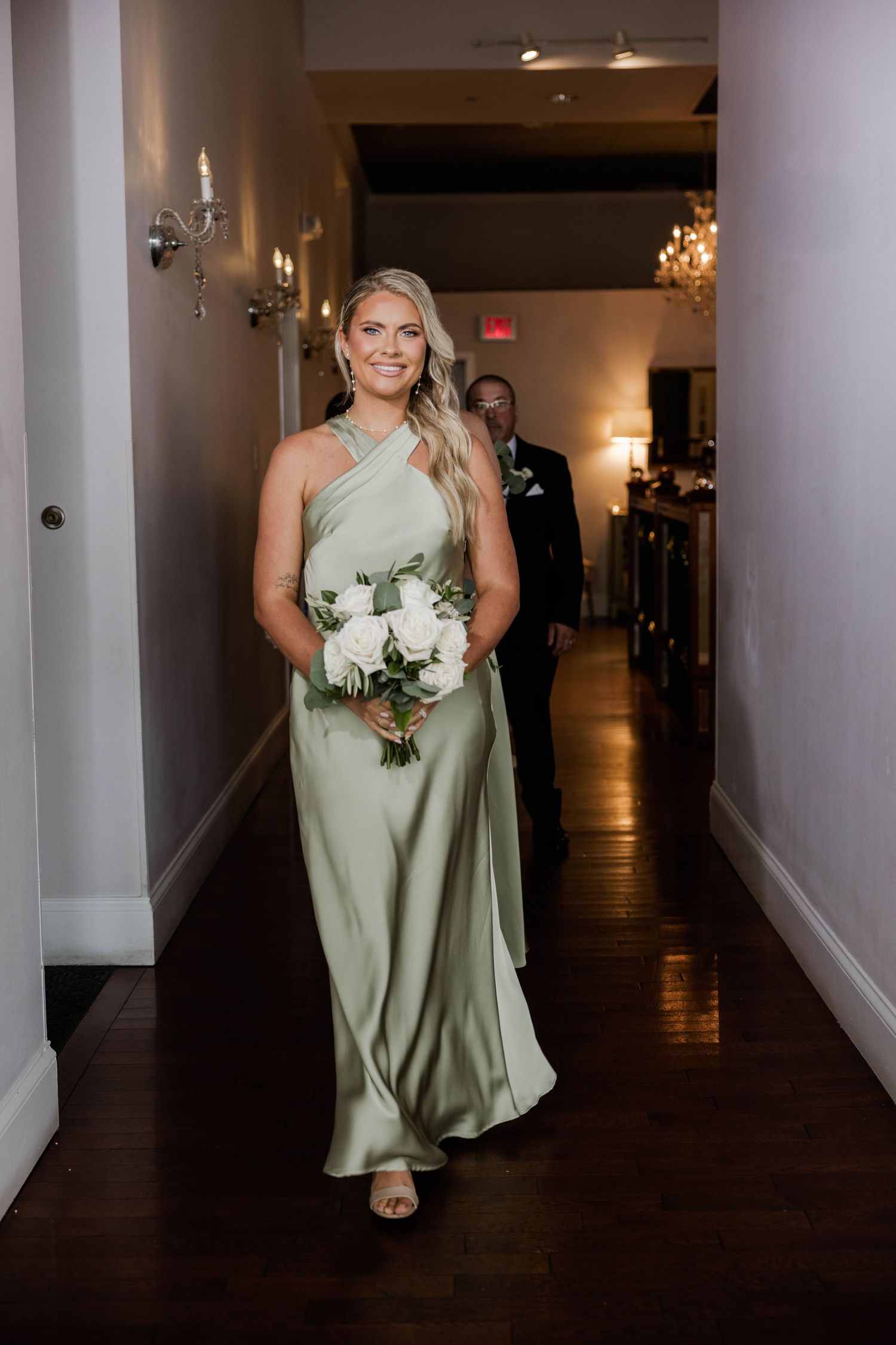 Bridesmaid in sage green satin halter gown walks down hallway holding white rose bouquet at wedding.