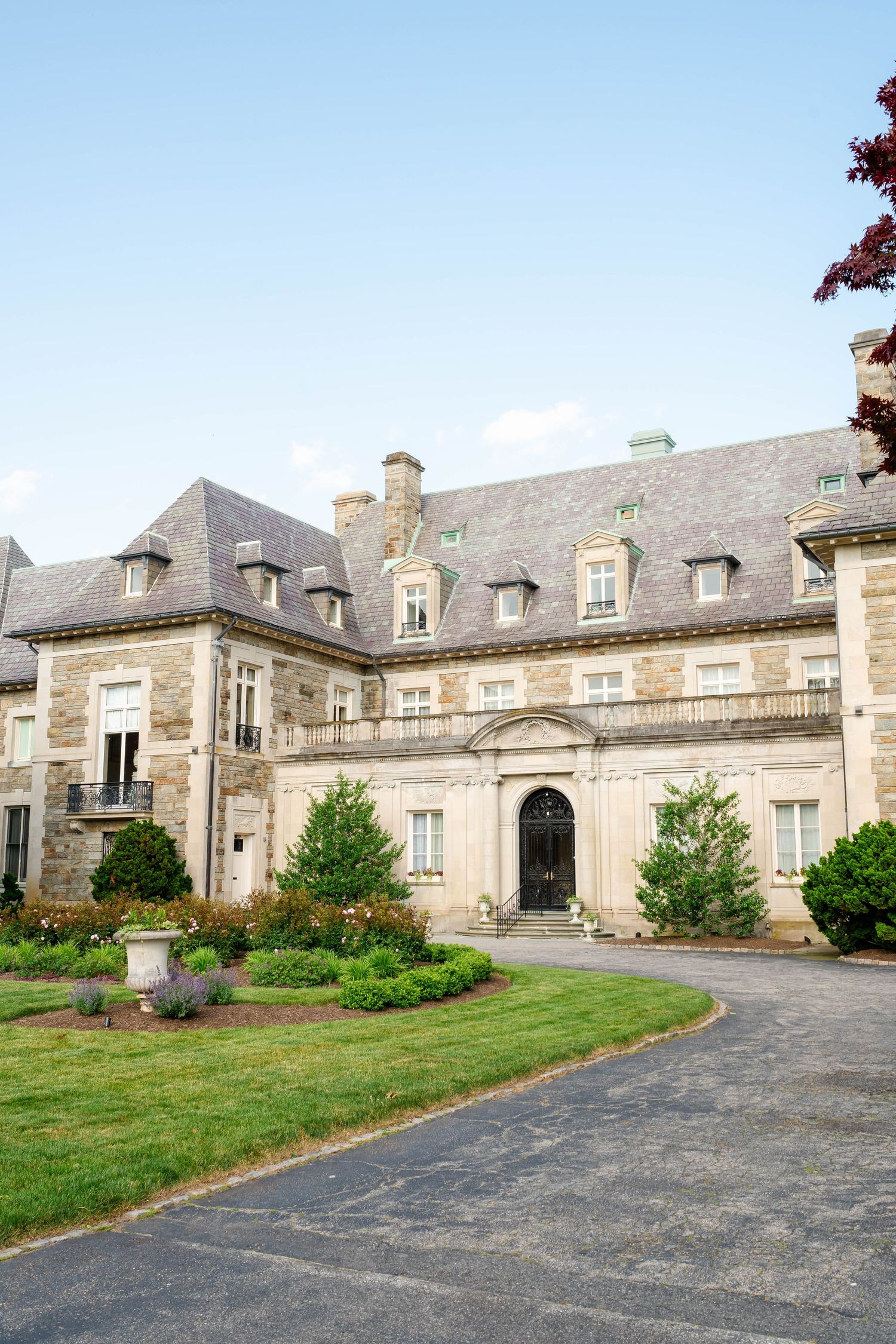 Grand stone manor estate with manicured gardens and circular driveway under blue sky.