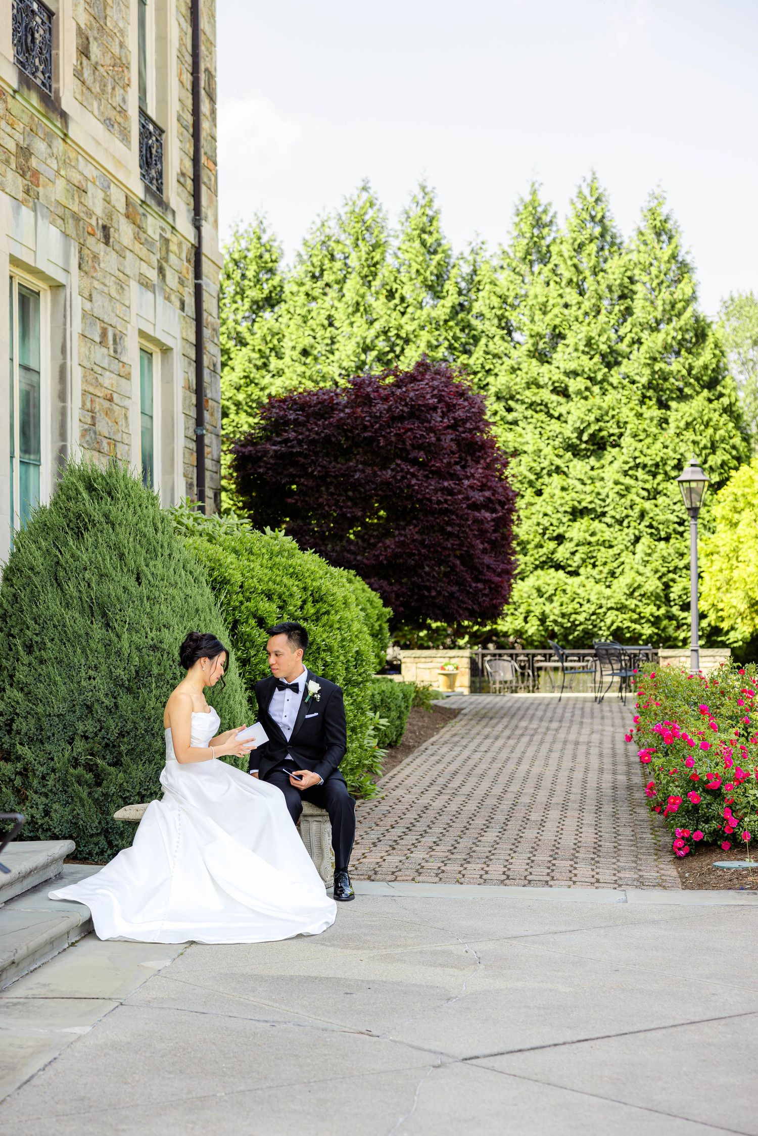 Bride and groom sit together outside stone building surrounded by lush green trees.