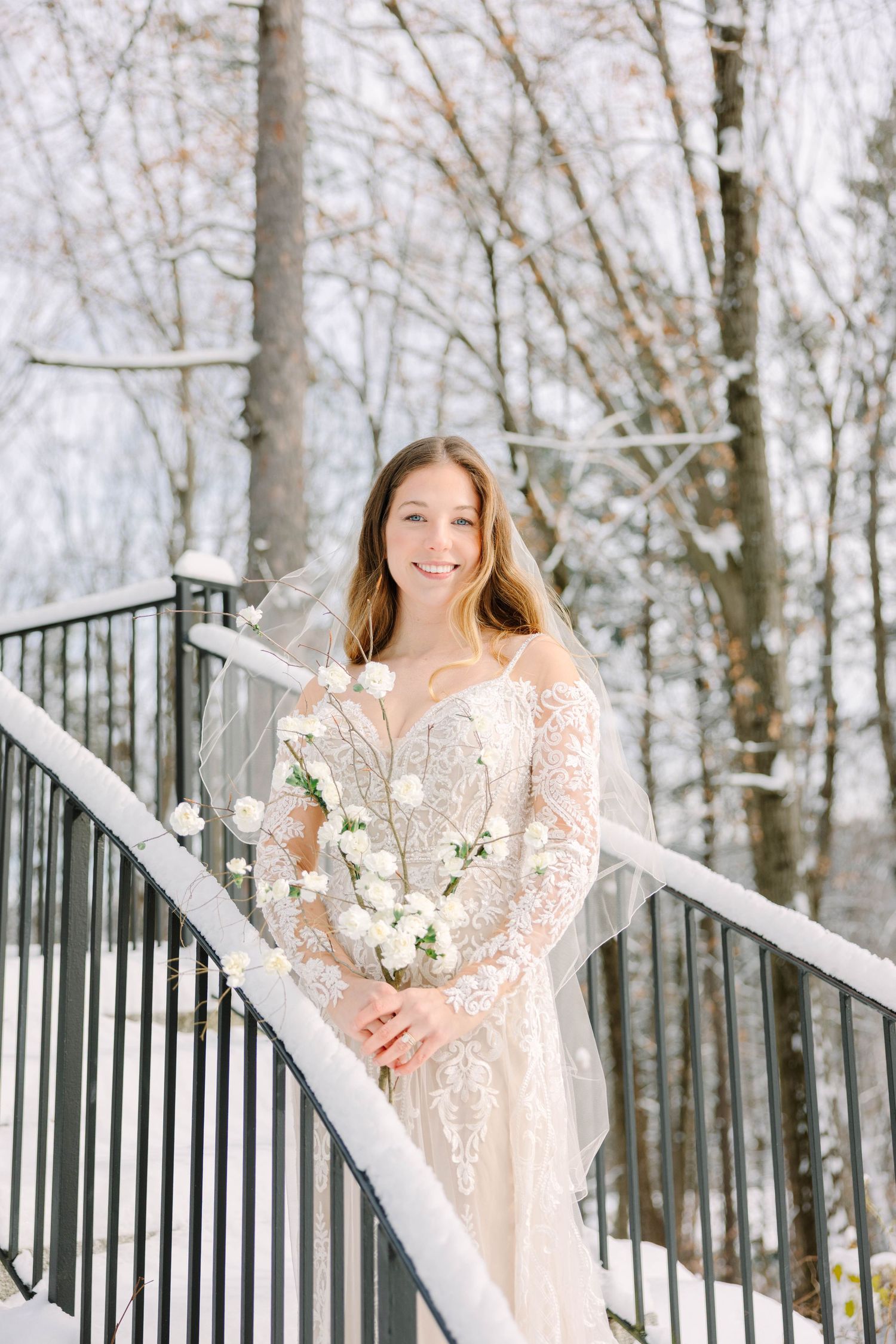 Smiling bride in lace gown and veil poses on snow-covered railing with white floral bouquet outdoors.