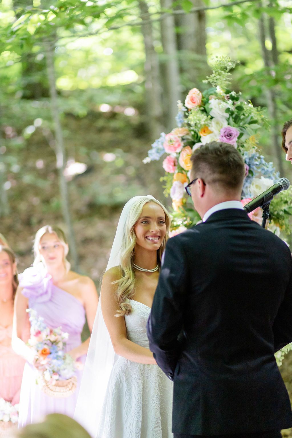 Radiant blonde bride smiling at groom during outdoor woodland wedding ceremony with colorful floral arch.