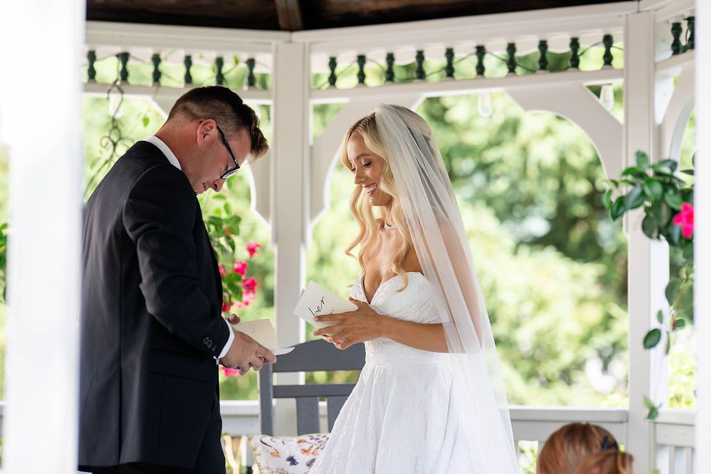 Bride and groom exchanging vows in a white gazebo surrounded by lush greenery and pink flowers.
