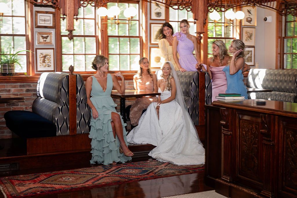 Bride in white gown sits with bridesmaids in colorful dresses in an elegant wood-paneled room.