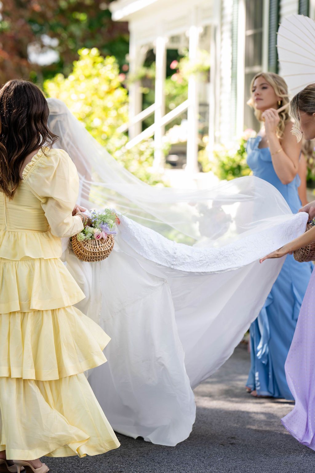Bridesmaids in yellow and blue dresses help arrange bride's flowing white veil and train outdoors.