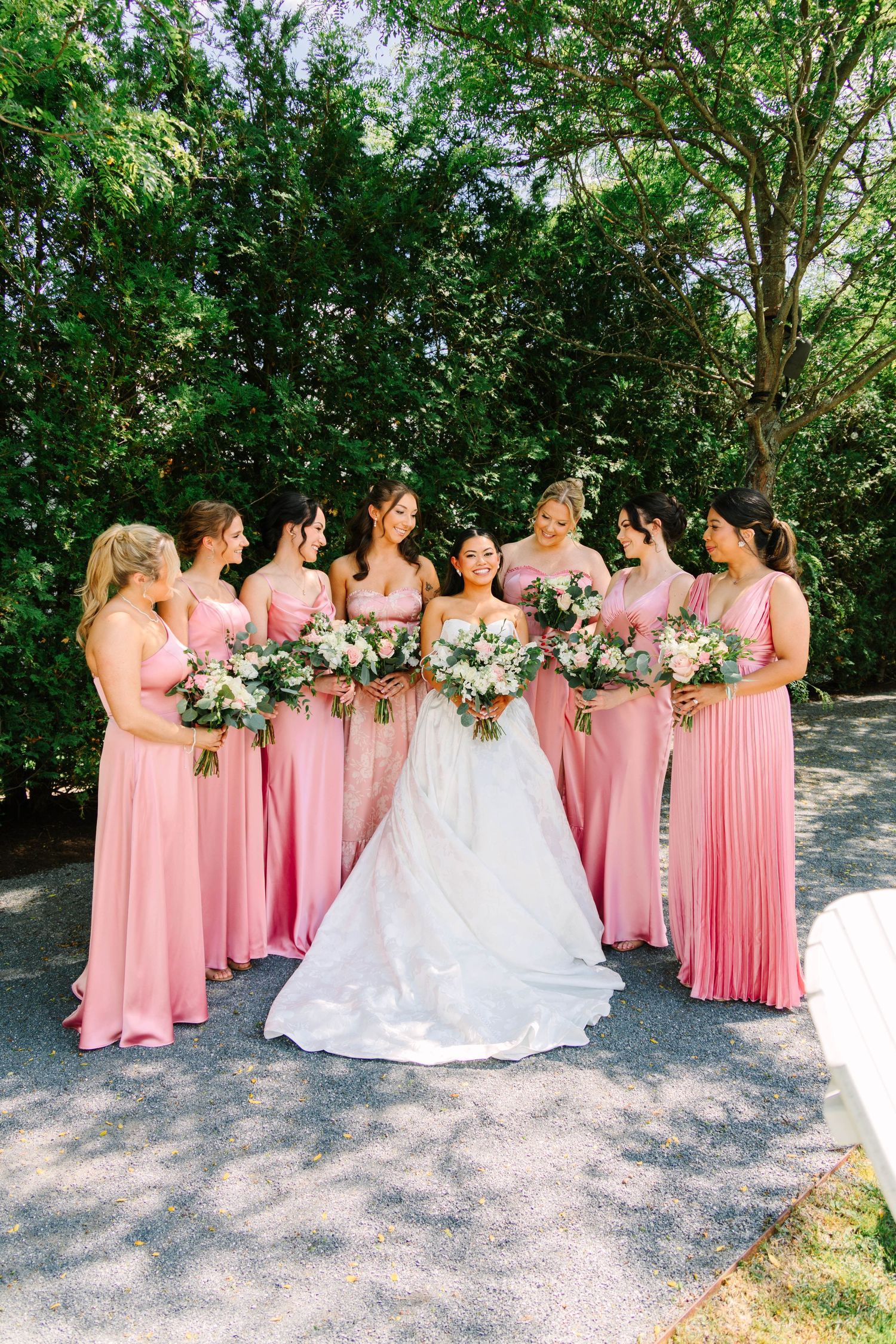 Bride in white gown surrounded by seven bridesmaids in pink dresses holding bouquets outdoors.