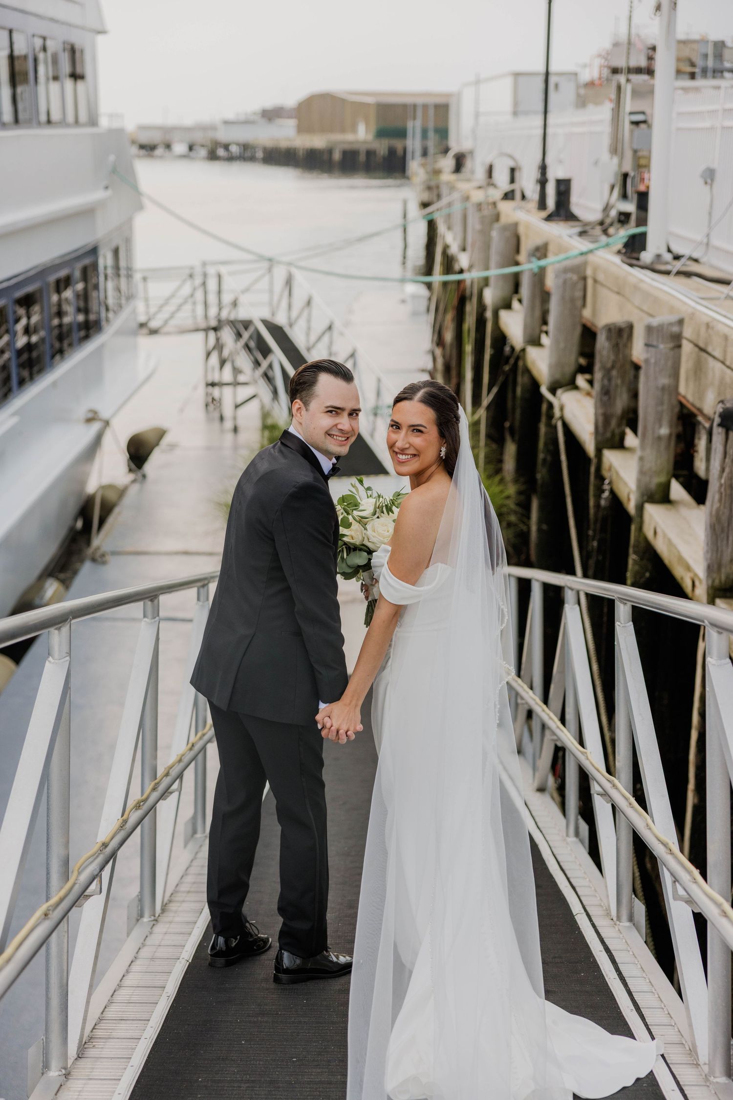 Bride and groom holding hands on a marina dock, smiling at camera, bride wearing elegant white gown with veil.