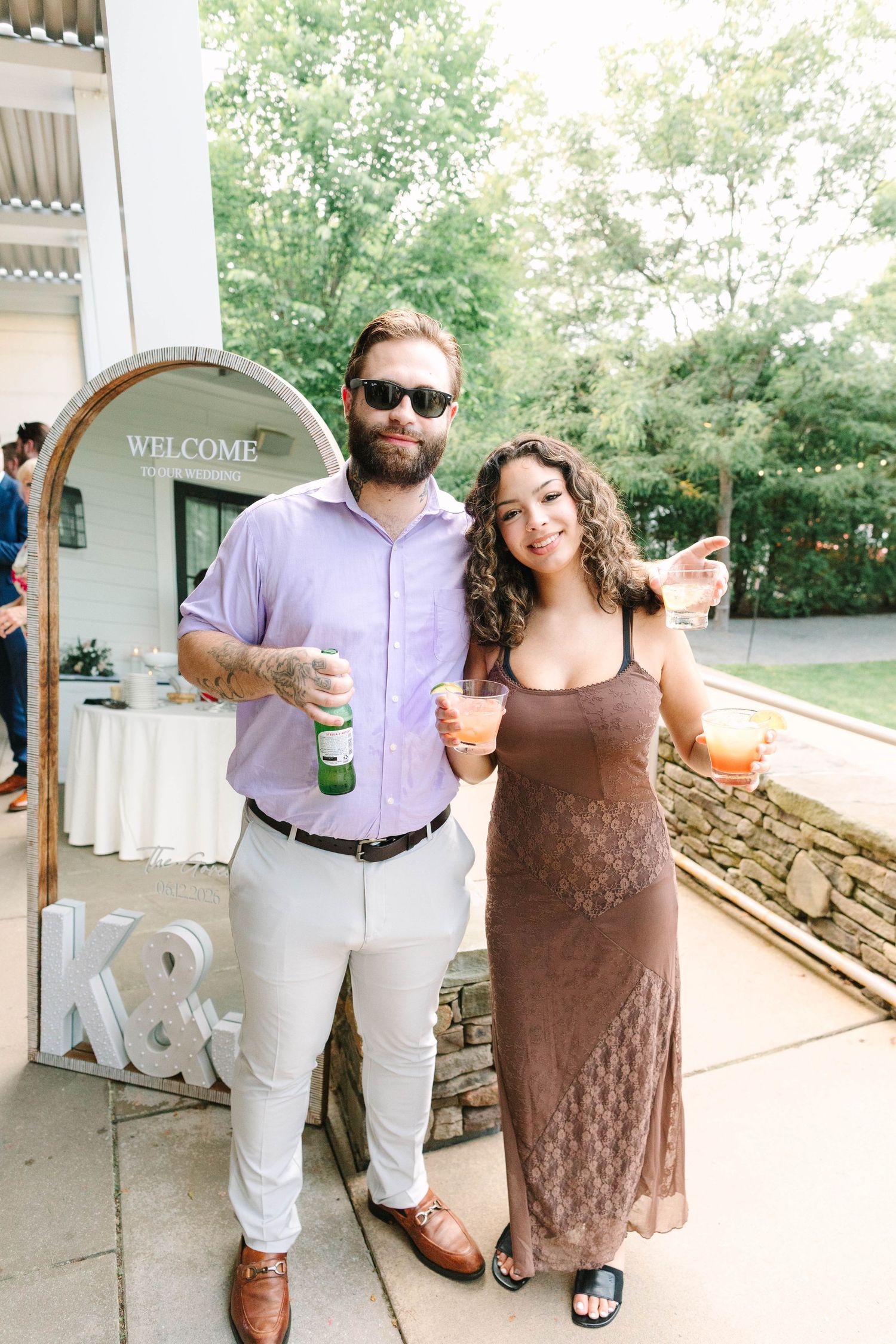 Wedding guests pose with drinks in front of a K&J monogram welcome sign at reception.