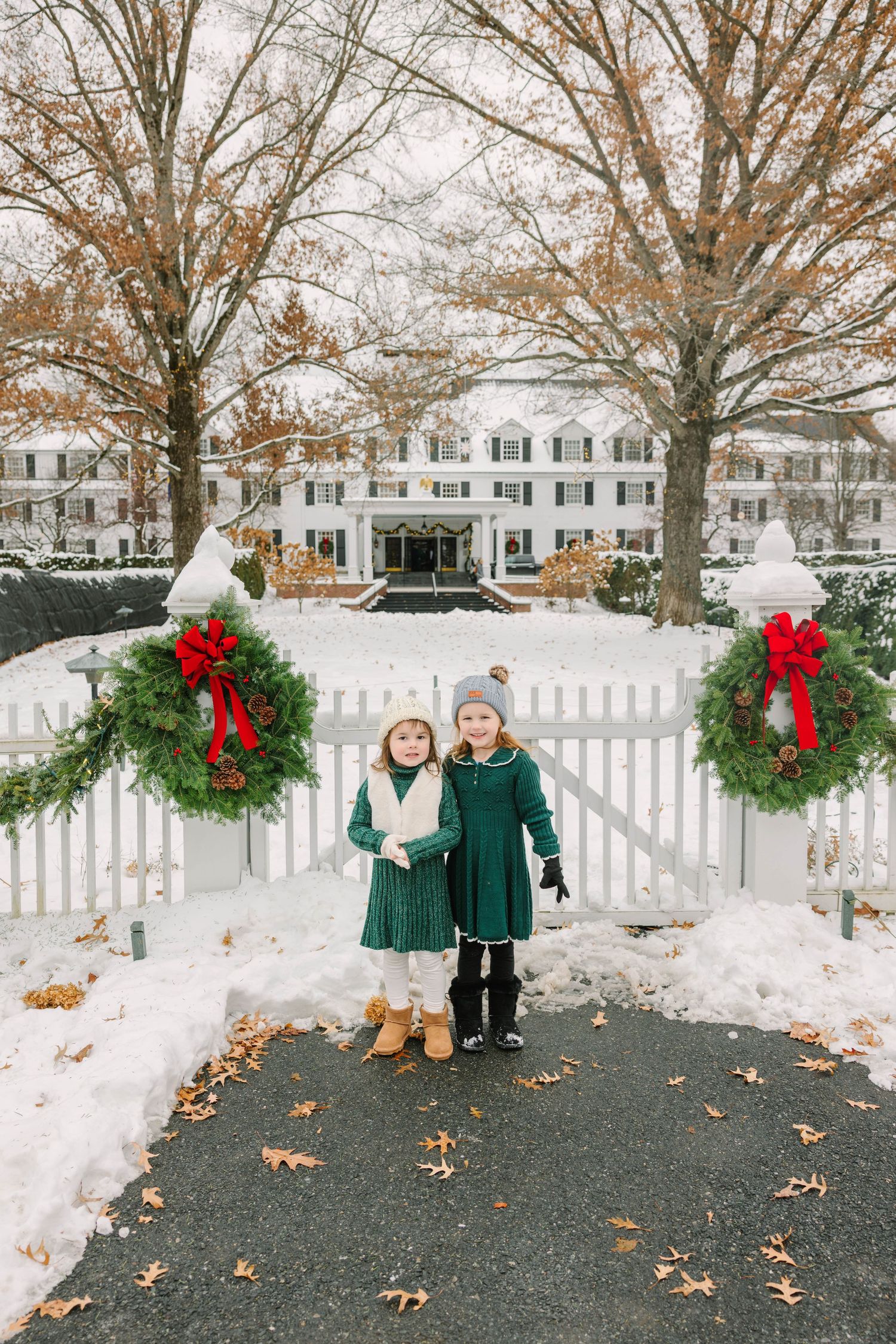 Two little girls in green dresses pose at a white fence gate adorned with Christmas wreaths and red bows in the snow.