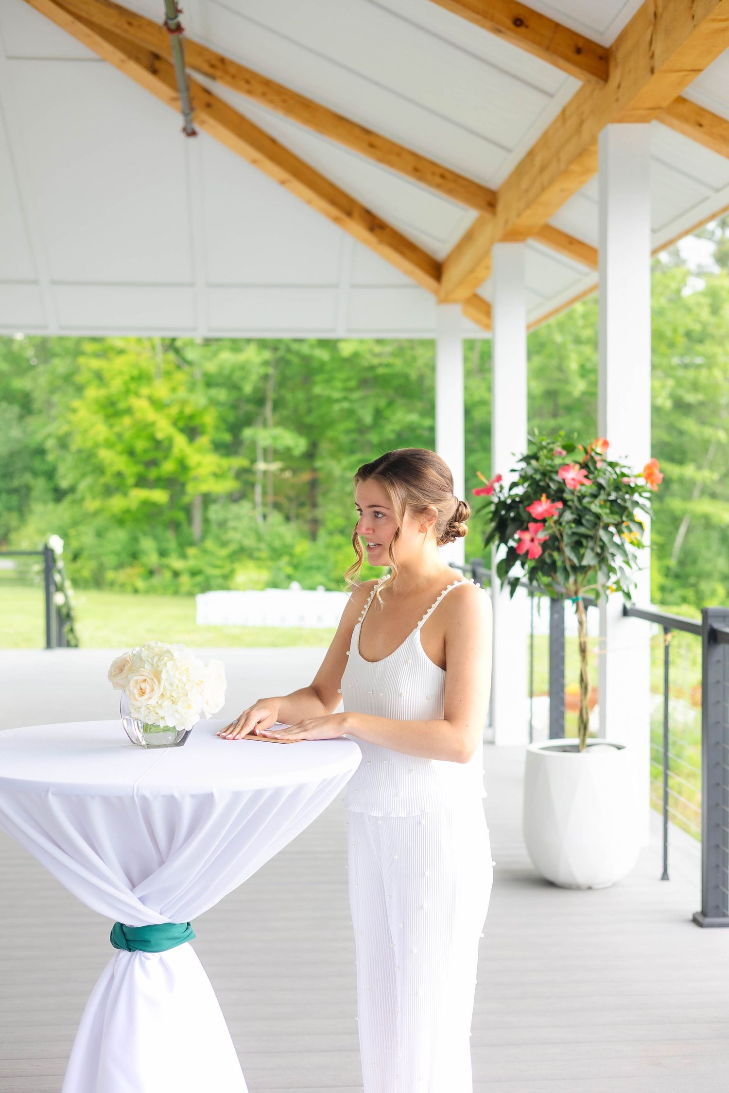 Elegant bride in a white dress standing thoughtfully at a draped cocktail table on a lush outdoor pavilion.