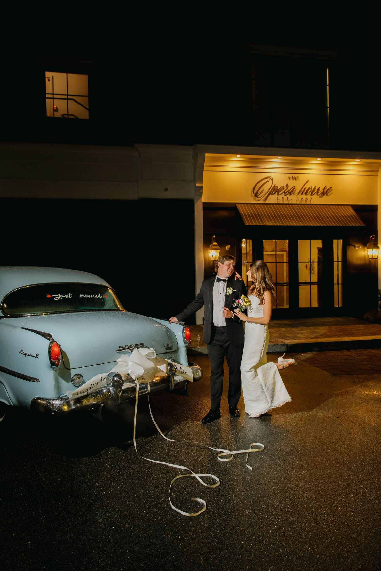 Bride and groom pose beside a vintage light blue car outside The Opera House venue at night.