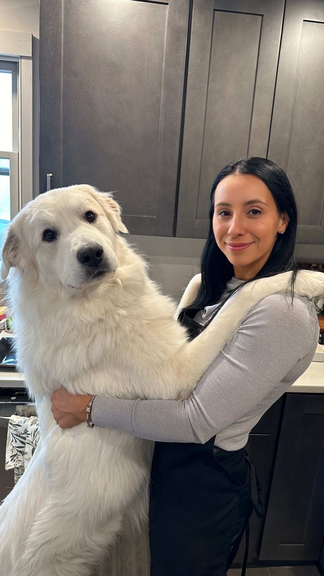 Woman smiling and hugging a large fluffy white Great Pyrenees dog in a modern kitchen.