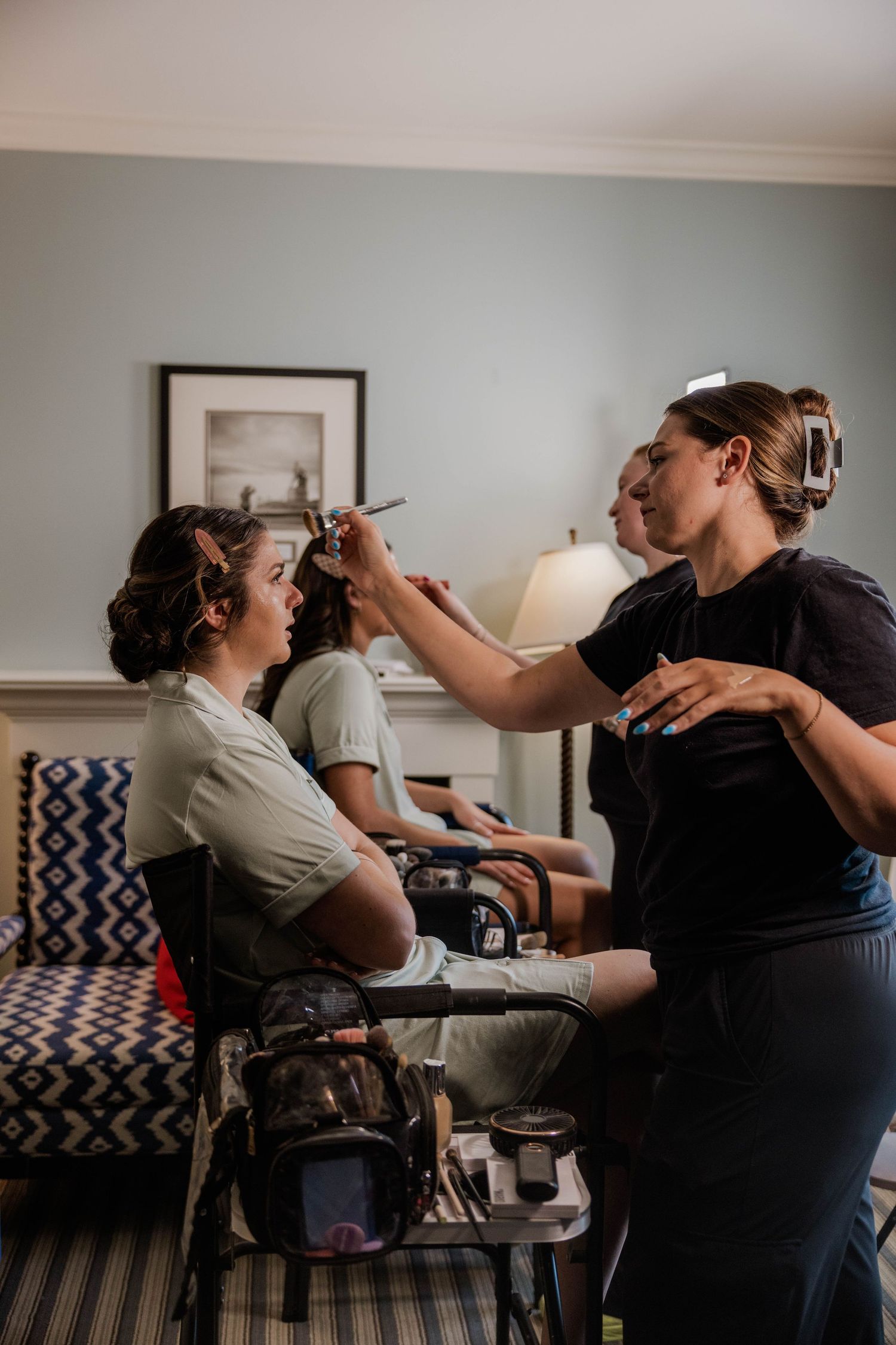 Makeup artist applying makeup to a bride while another woman sits in the background in a hotel room.