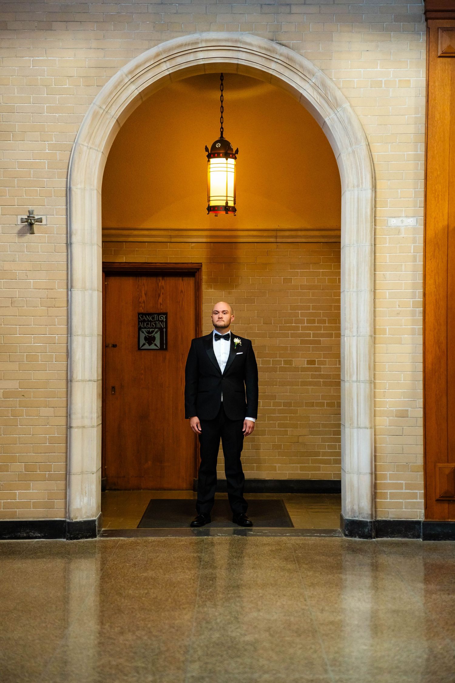 Groom in black tuxedo standing under a stone arch with hanging lantern in historic venue.