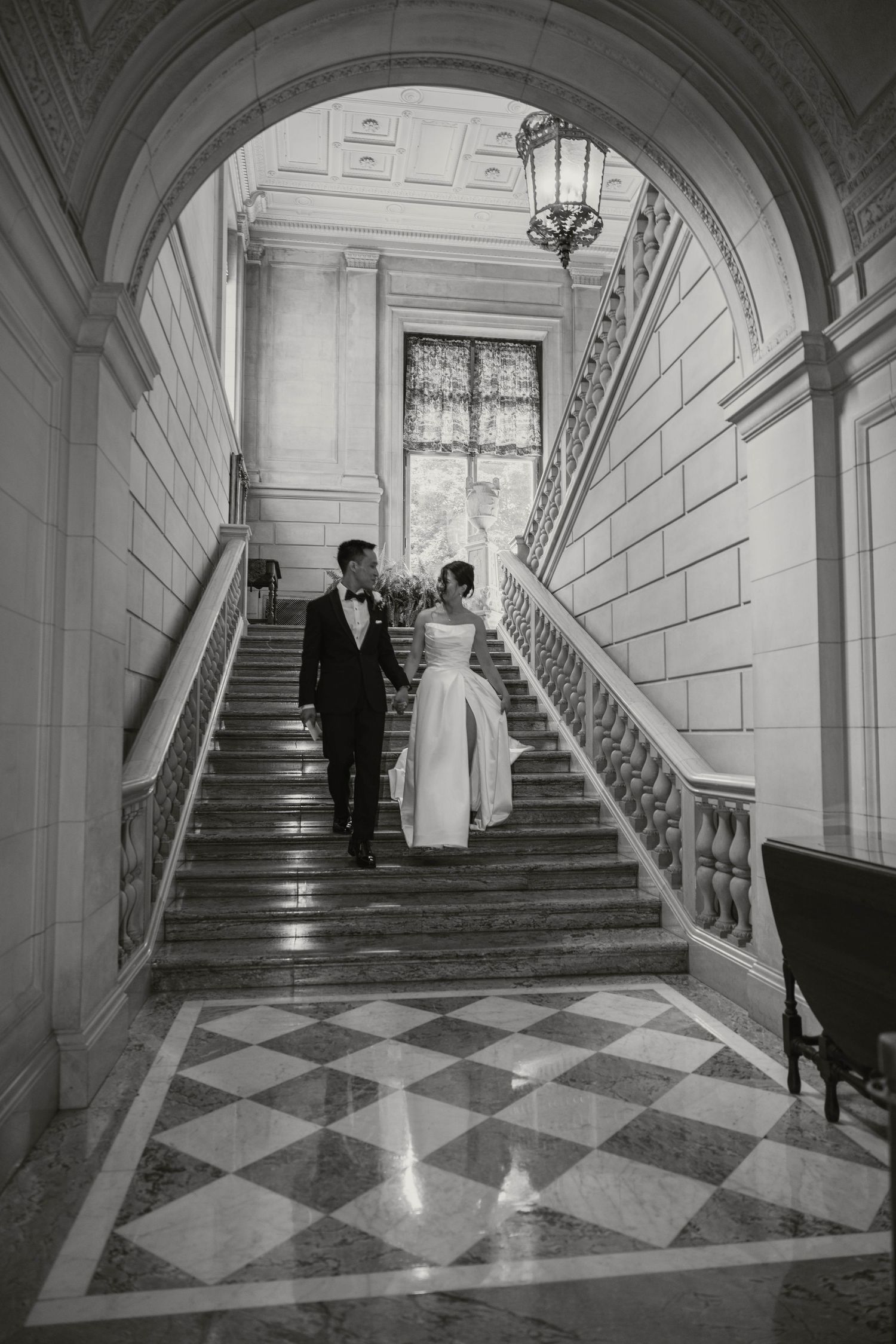 Black and white photo of bride and groom holding hands walking down grand marble staircase with arched entry.