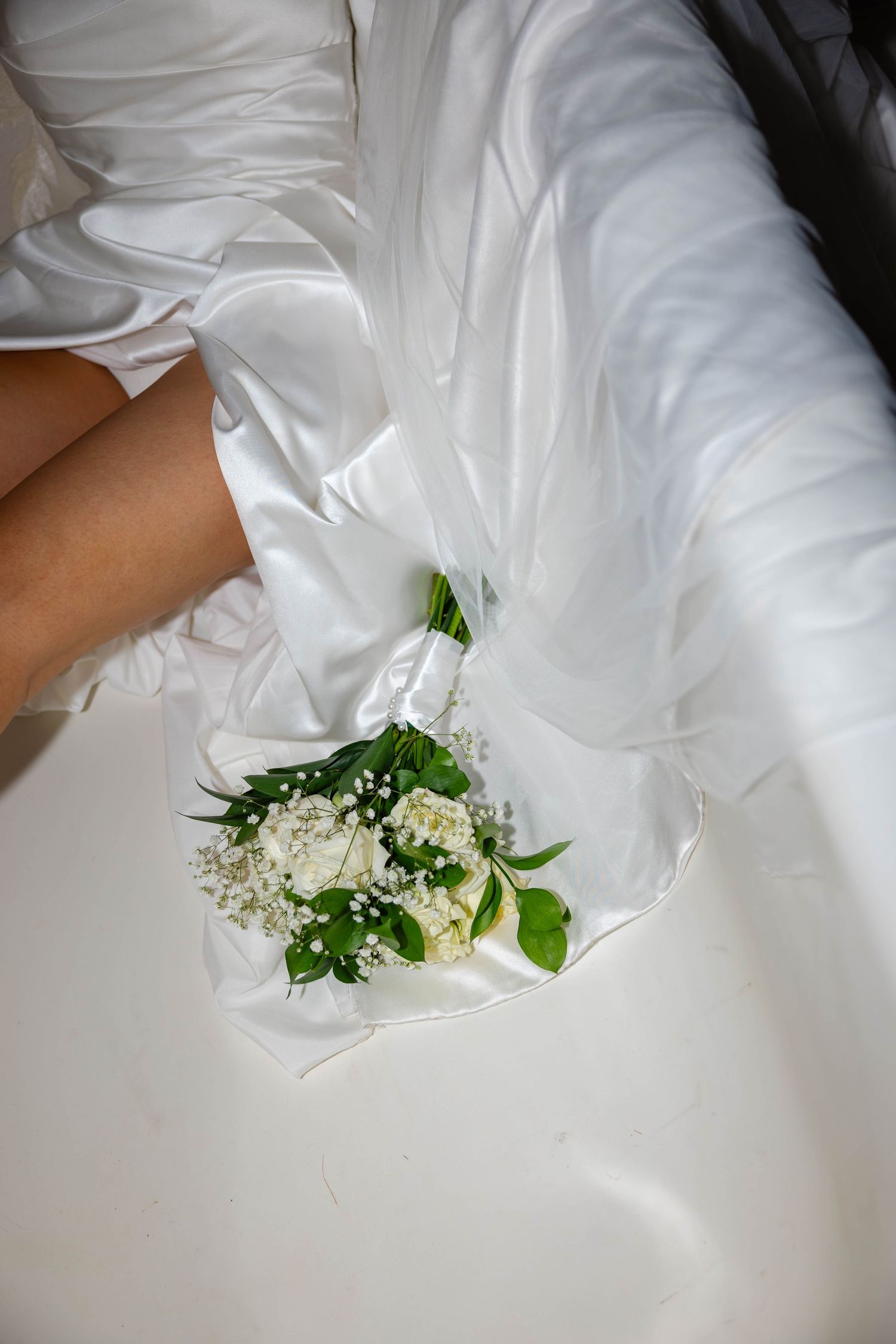 Close-up of bride in white satin gown holding a delicate bouquet of white roses and baby's breath in a bathtub.