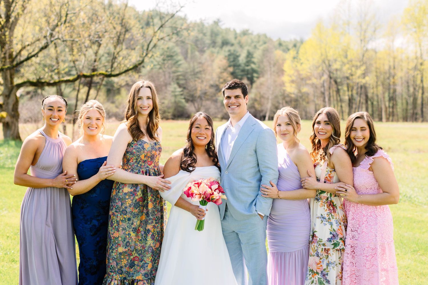 Bride and groom posing with bridesmaids in colorful dresses in a lush green outdoor setting.