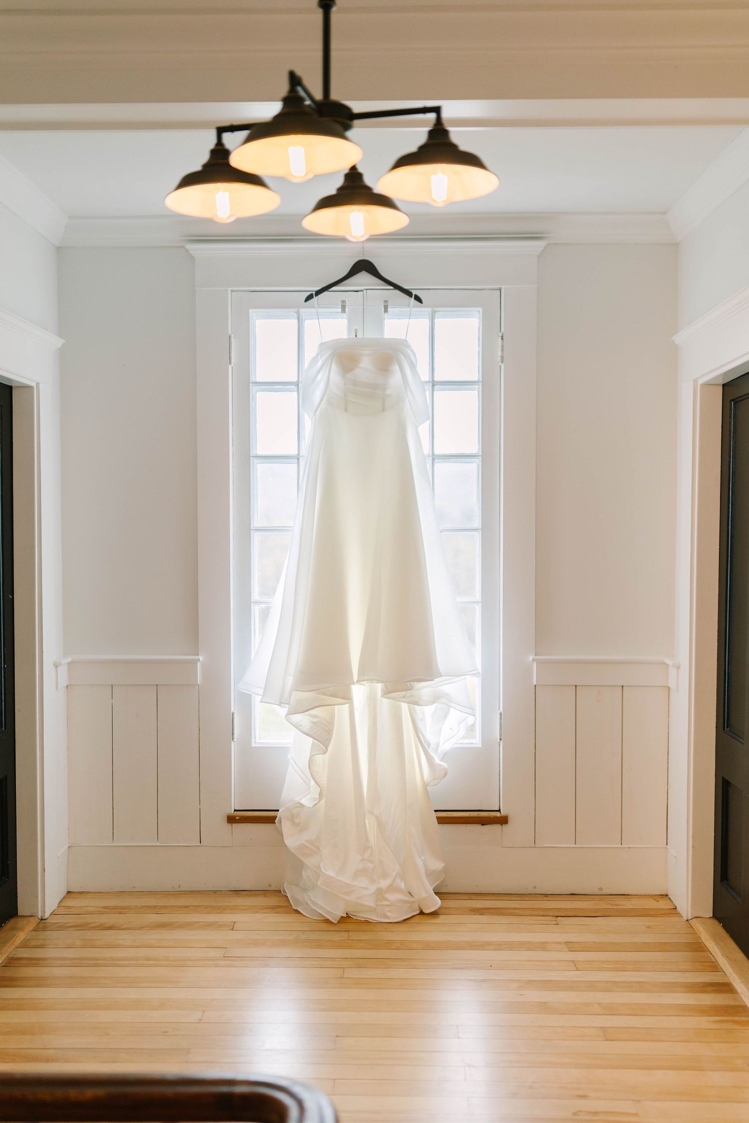 Elegant white wedding dress hanging from black chandelier in front of white paneled window in bright airy room.