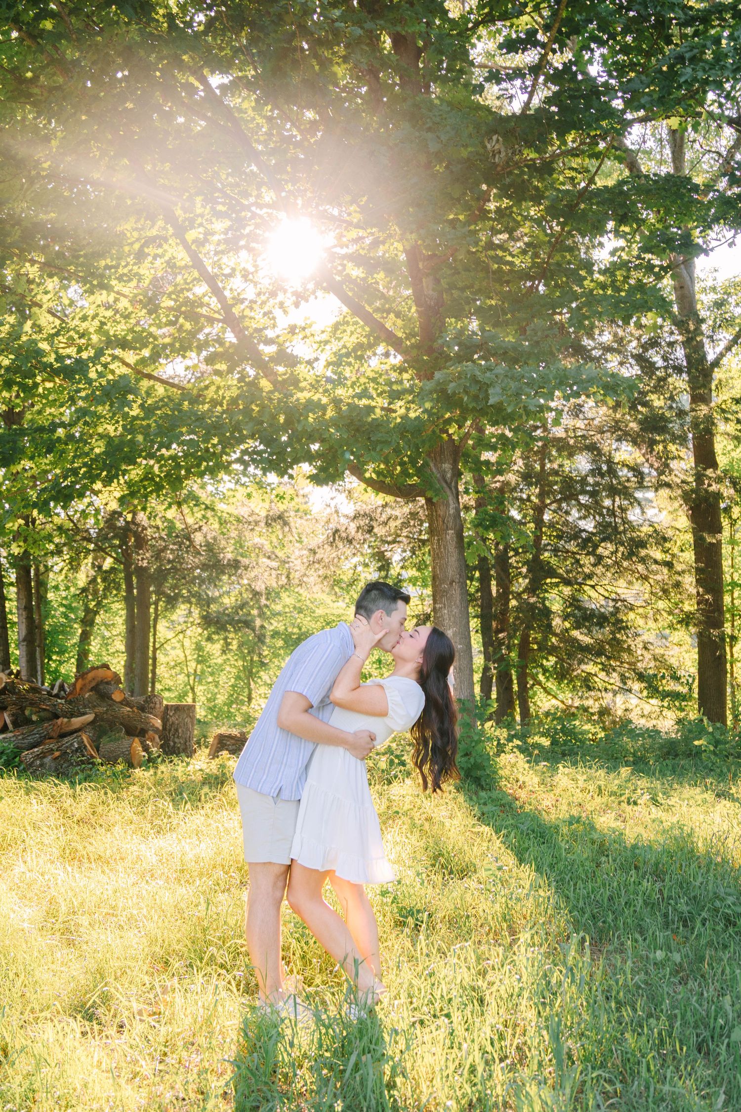 Couple sharing a romantic kiss in a sunlit forest clearing with golden backlight filtering through green trees.