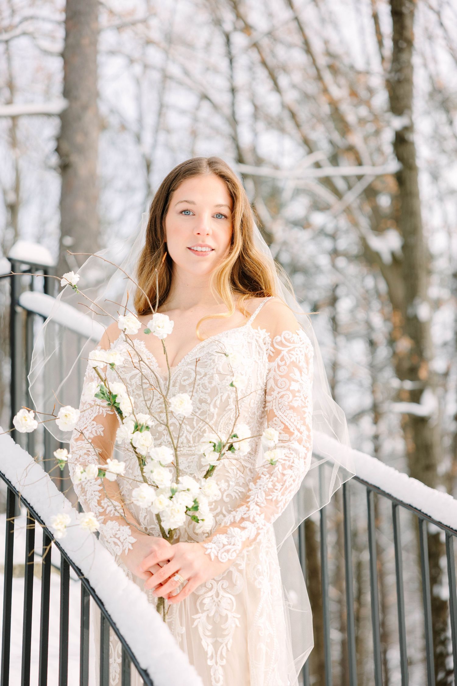 Bride in lace wedding dress and veil poses on snowy staircase railing with white bouquet.