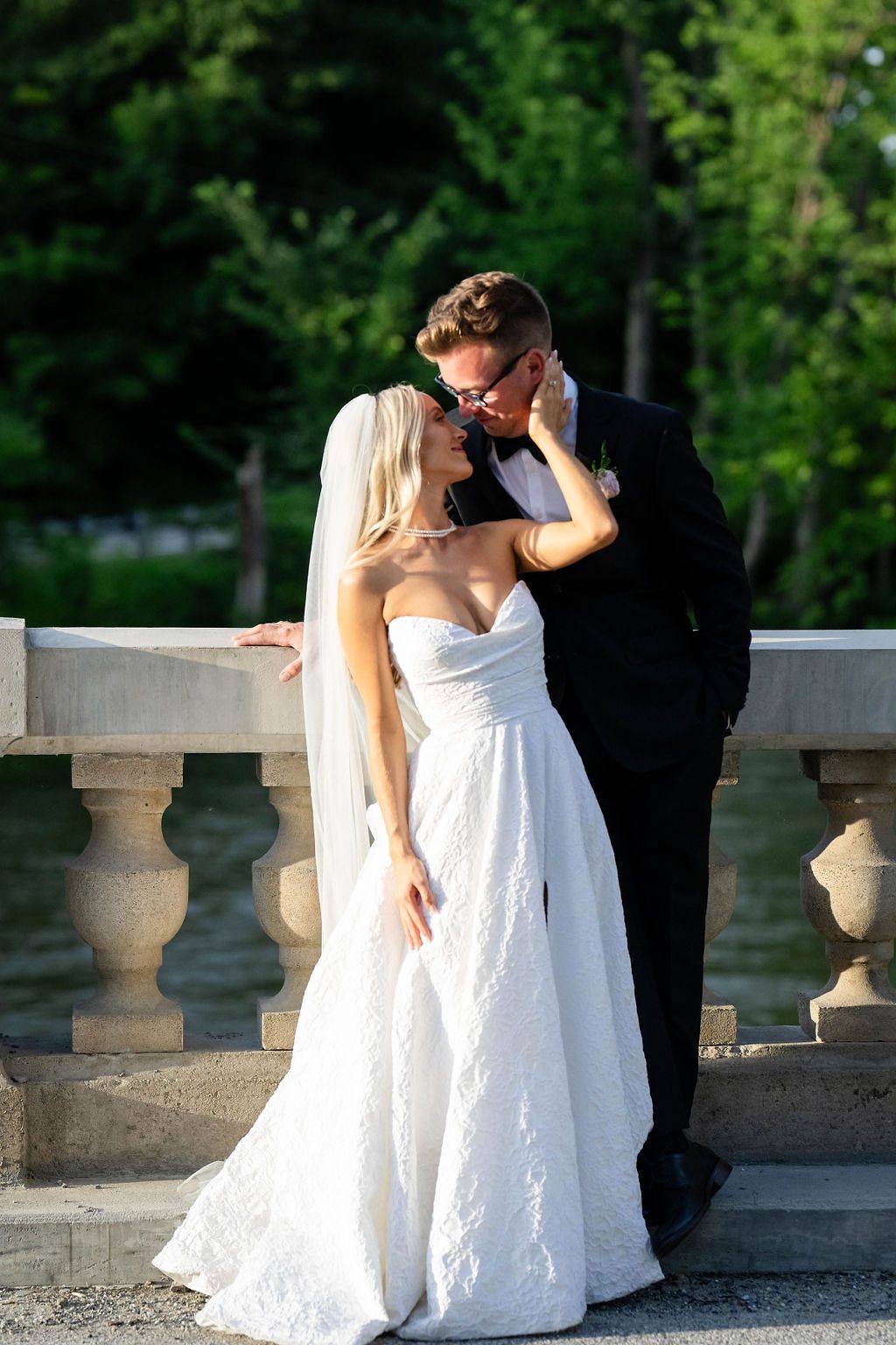 Bride and groom sharing a romantic kiss on a stone balustrade bridge surrounded by greenery near a lake.