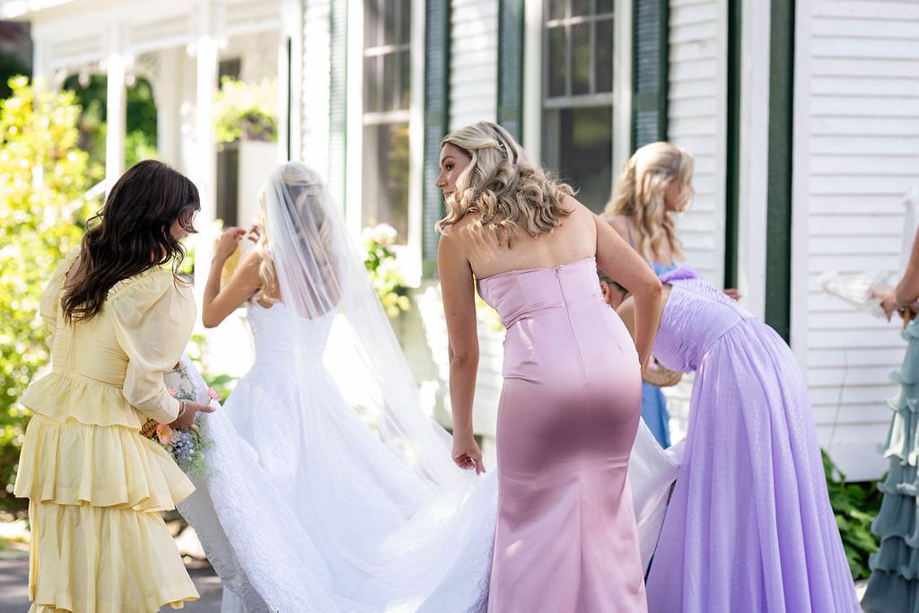 Bridesmaids in colorful gowns adjust bride's veil and train outside a white venue.