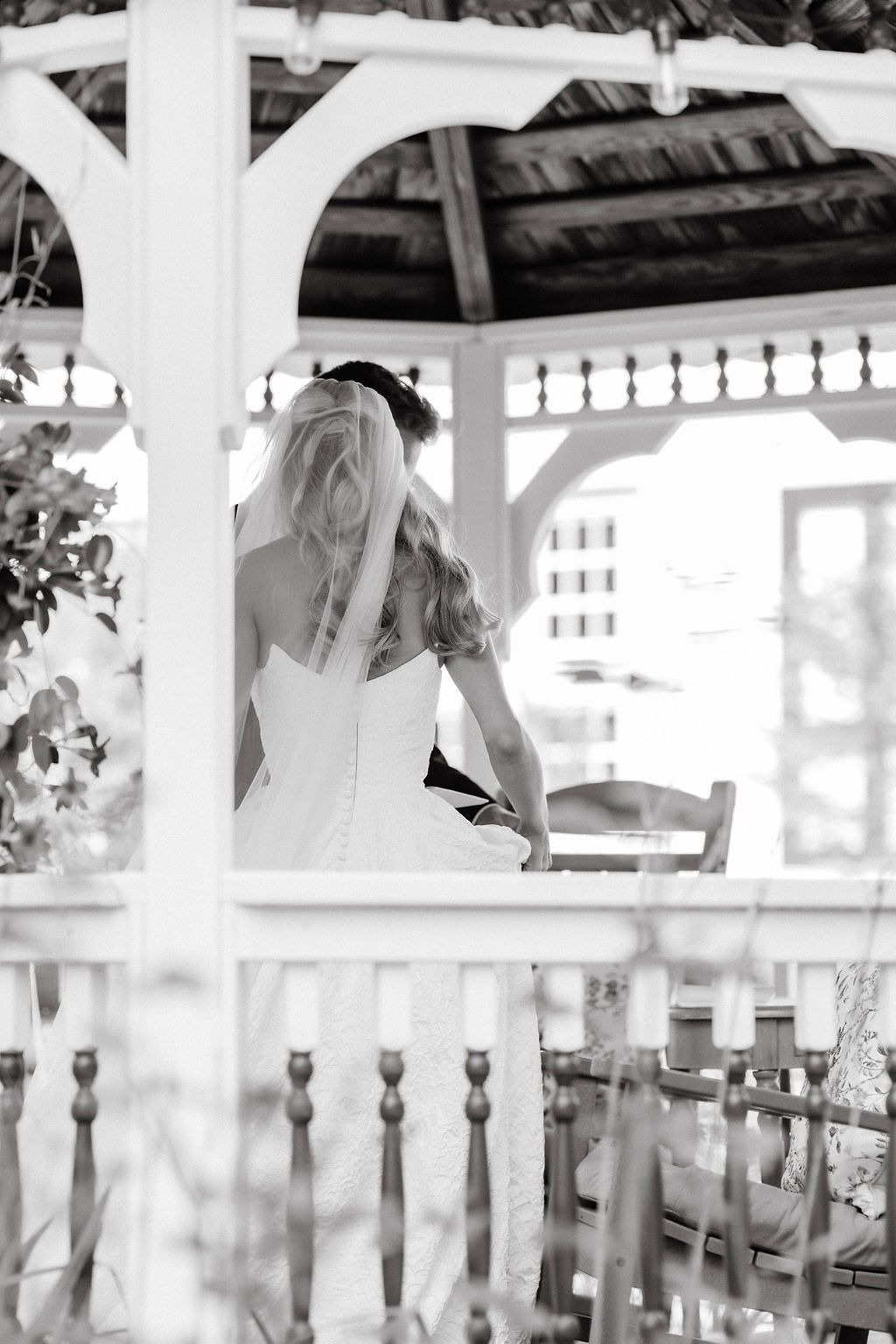 Black and white photo of bride in wedding gown and veil seated inside an elegant white gazebo.