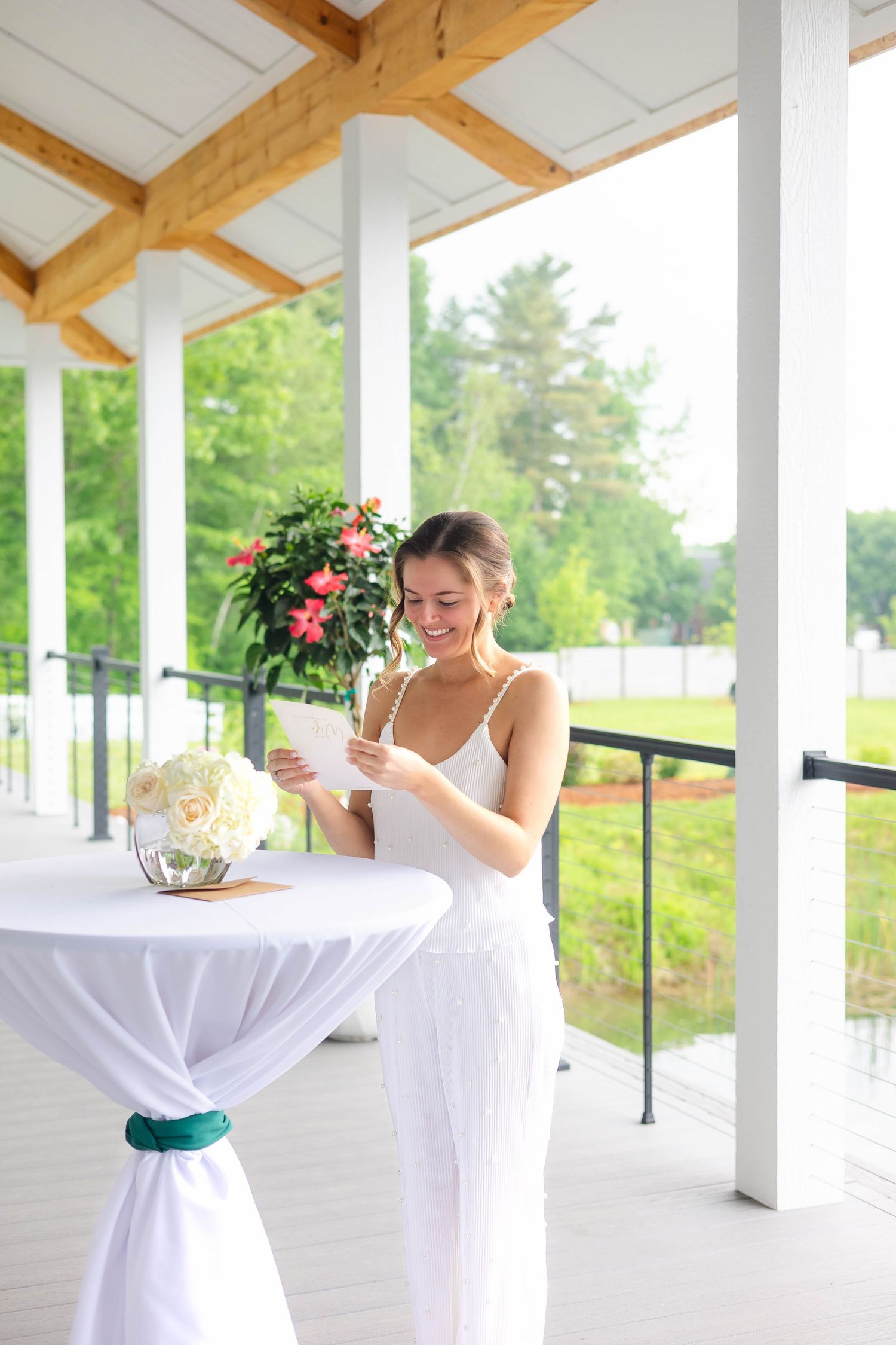 Smiling bride in a white dress reading a card at a draped cocktail table on a bright outdoor pavilion.