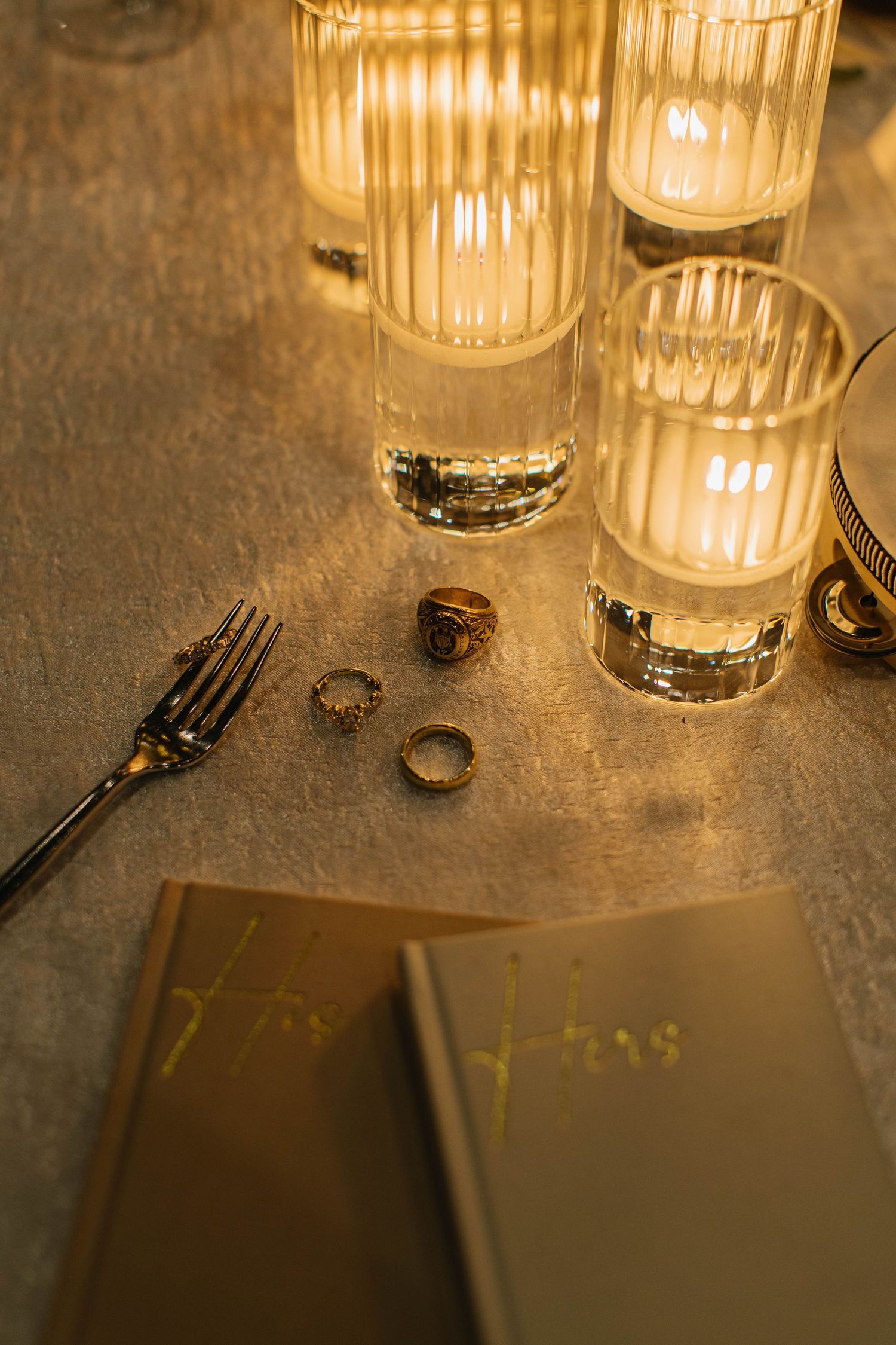 Elegant table setting with glowing ribbed candle holders, gold rings, a fork, and a golden menu on a metallic surface.