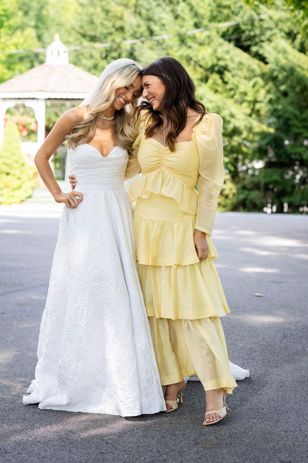Bride in strapless white gown laughs with bridesmaid in yellow ruffled dress near garden gazebo.