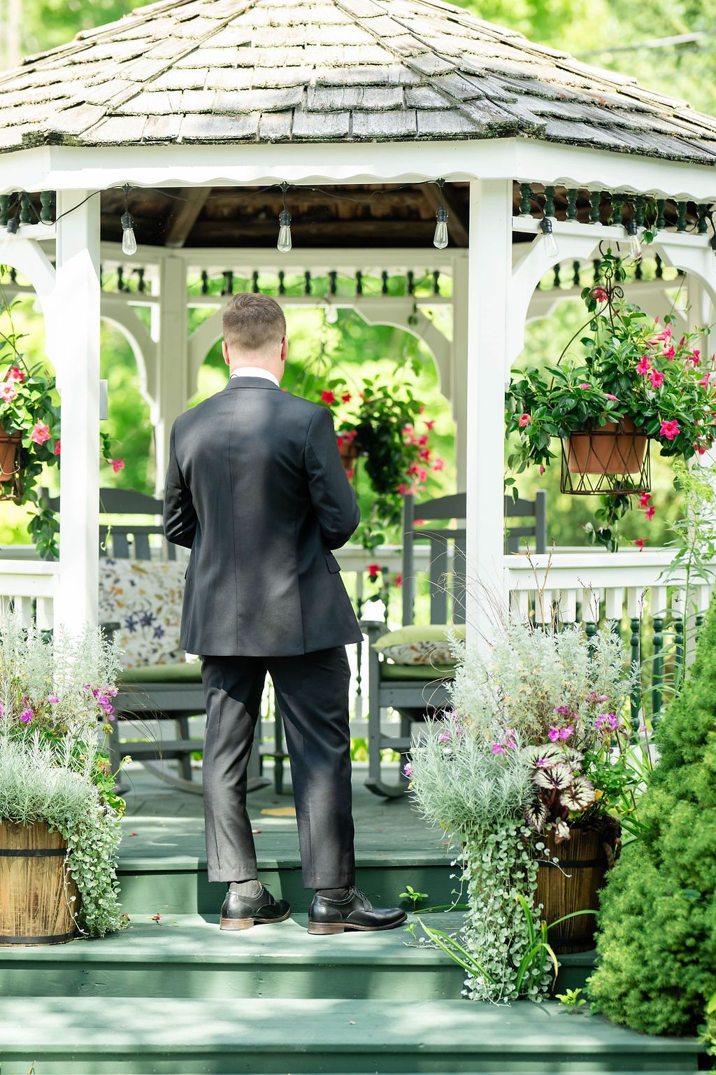 Man in dark suit standing at a white gazebo surrounded by lush flowering plants and hanging baskets in a garden.
