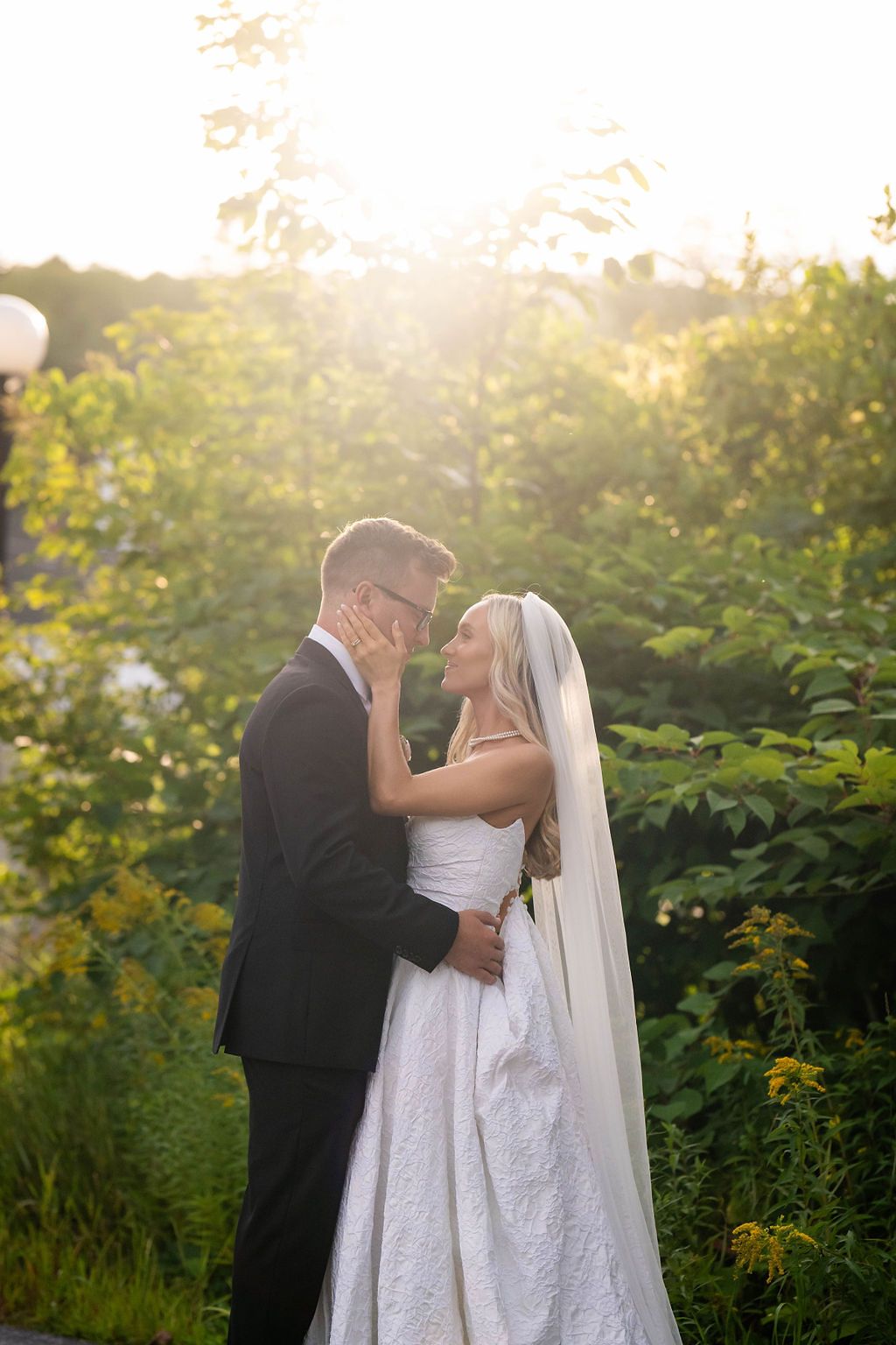 Bride and groom share a romantic moment surrounded by lush greenery in golden sunset light.