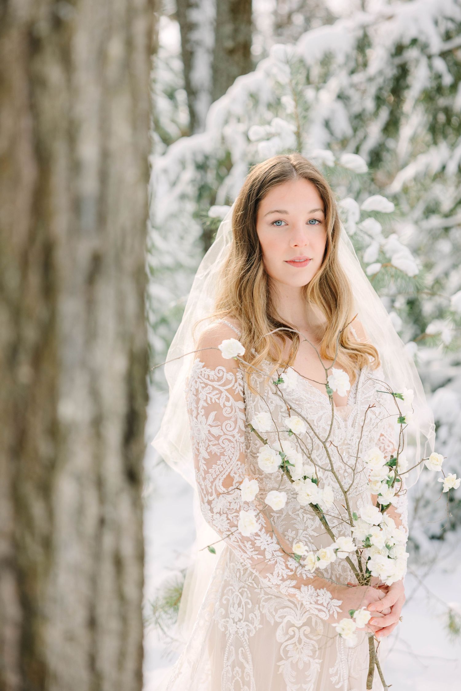Elegant bride wearing lace dress and veil holds delicate white flowers in a snowy winter forest.