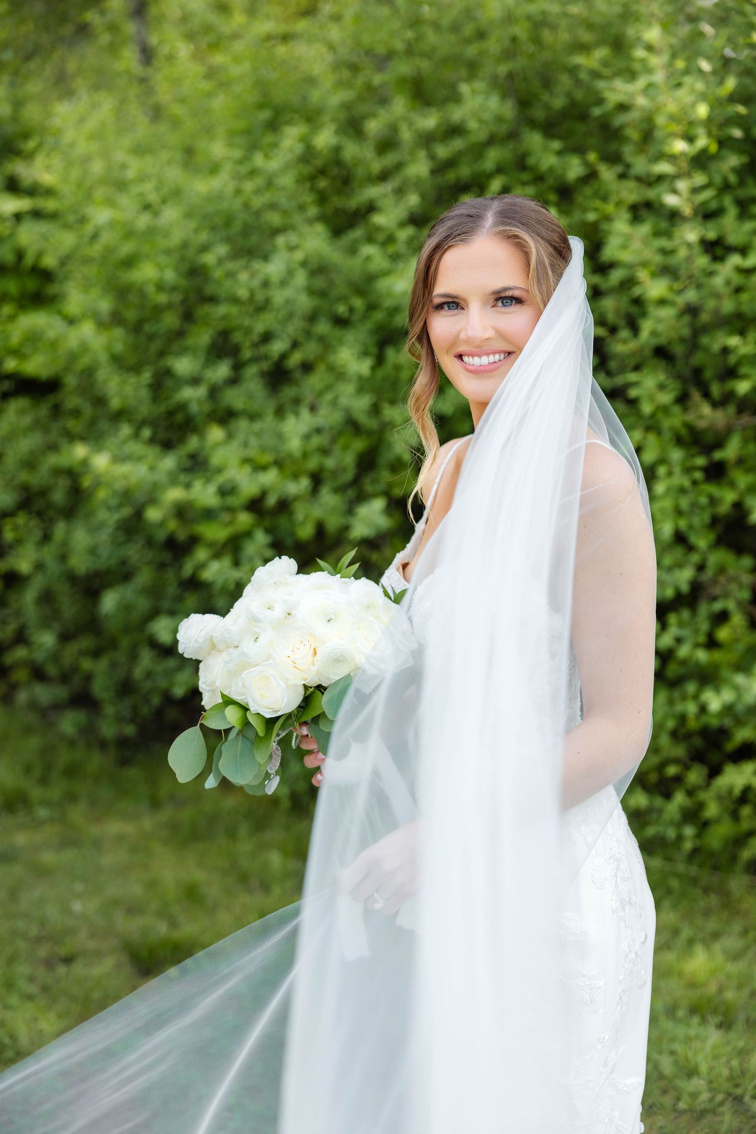 Smiling bride in lace gown holding white bouquet with flowing veil in lush green garden.