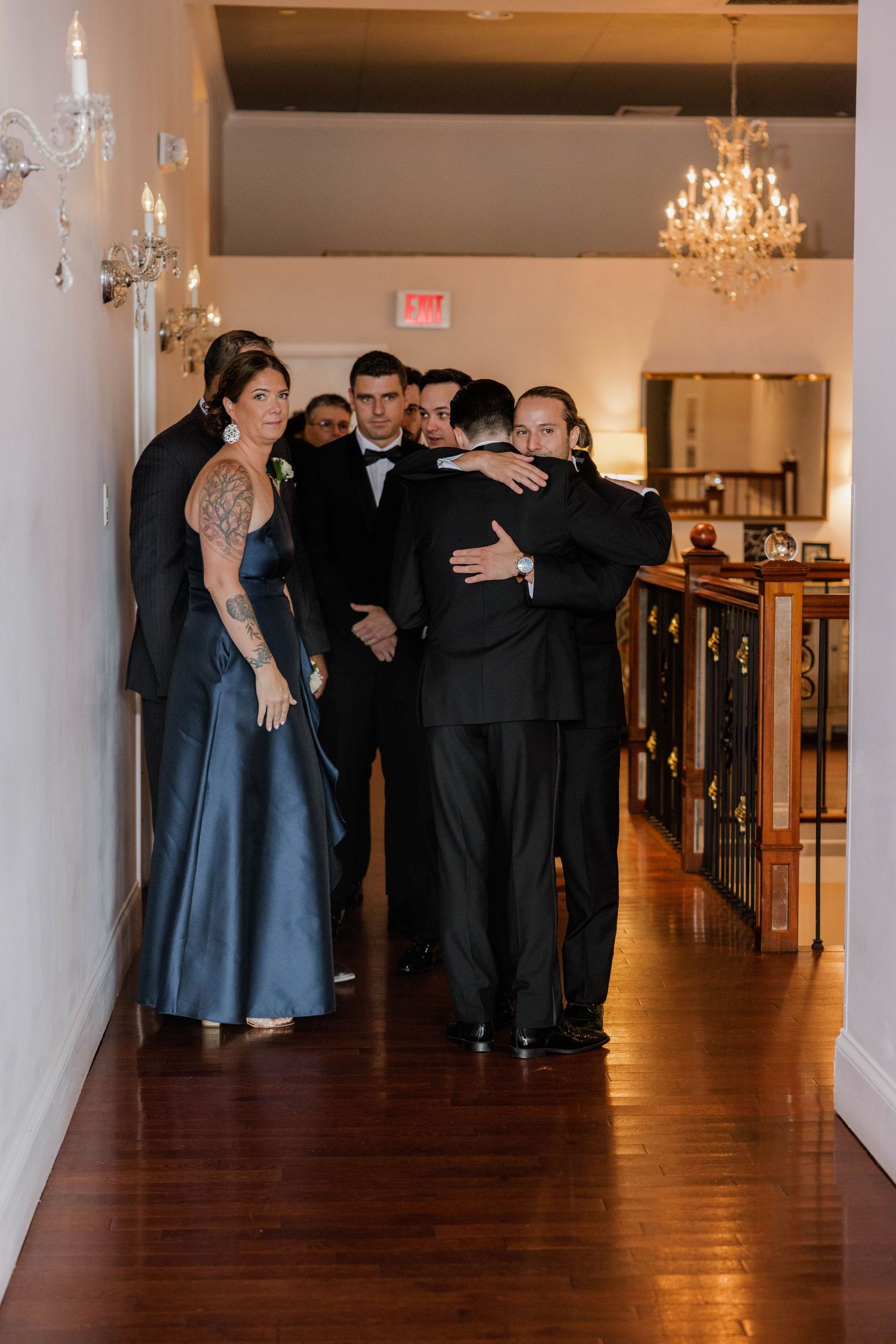 Wedding party in formal attire sharing an emotional embrace in a hallway.