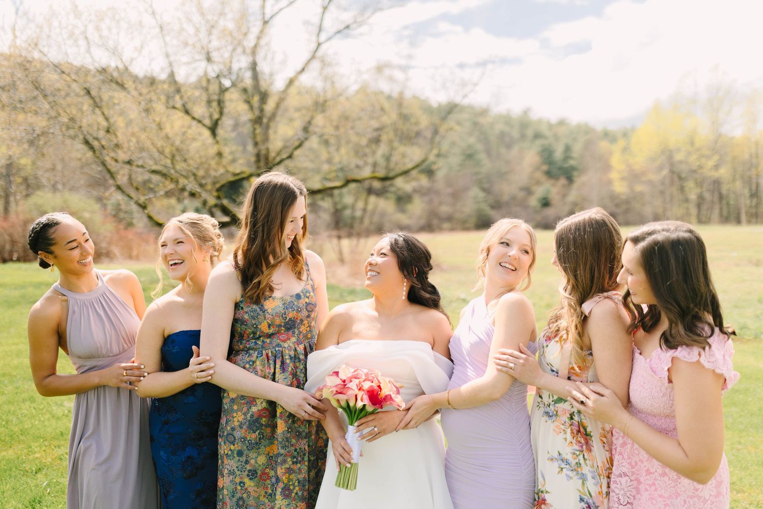 Bride with bouquet laughing with five bridesmaids in colorful dresses outdoors in a green field.