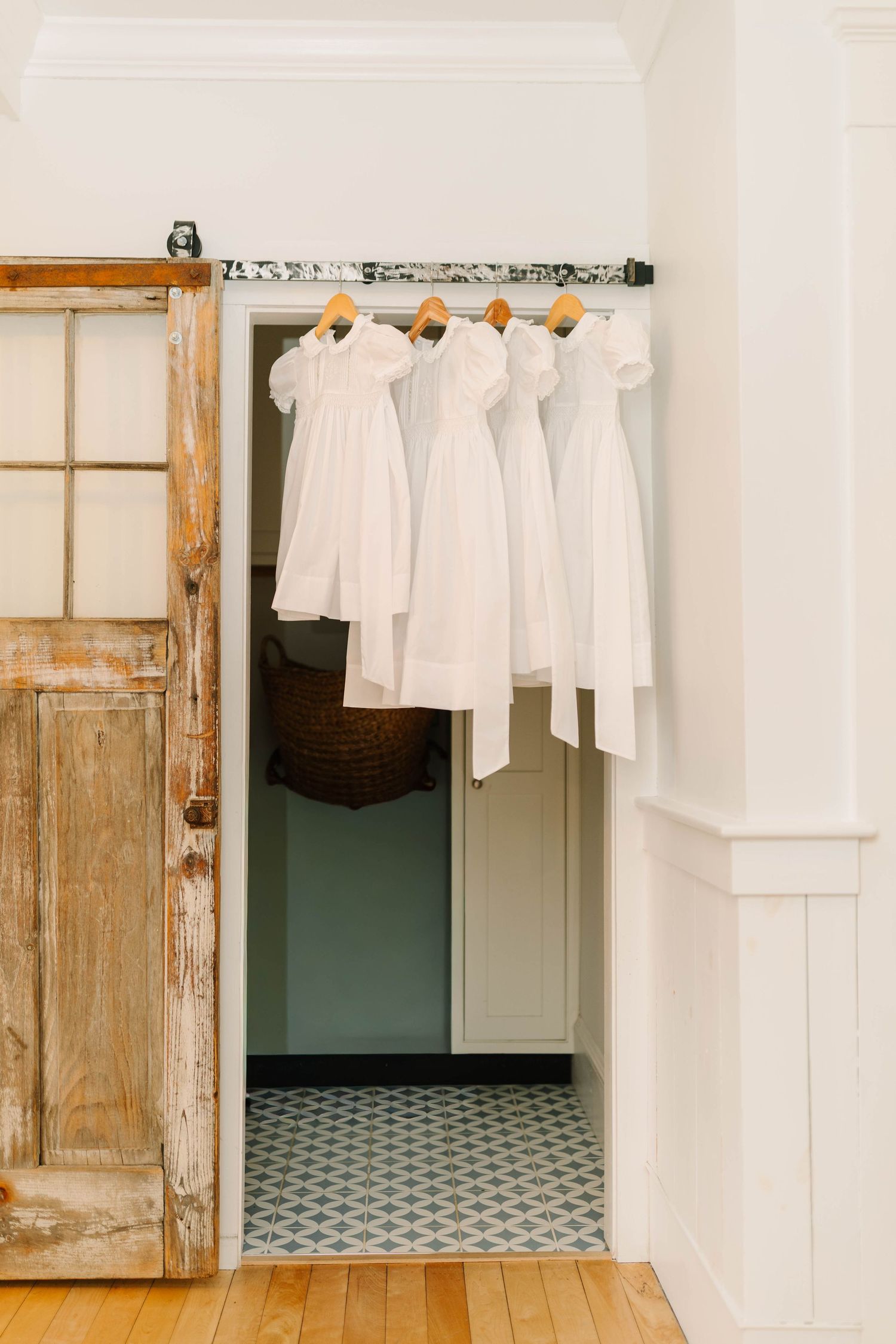 Four white dresses hanging on wooden hangers in a rustic closet with vintage door and patterned tile floor.