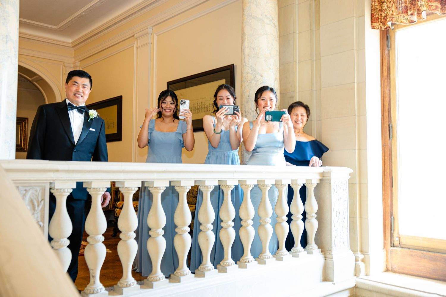 Wedding party poses on elegant balcony, bridesmaids in blue dresses taking photos, groom in tuxedo smiling at formal venue.