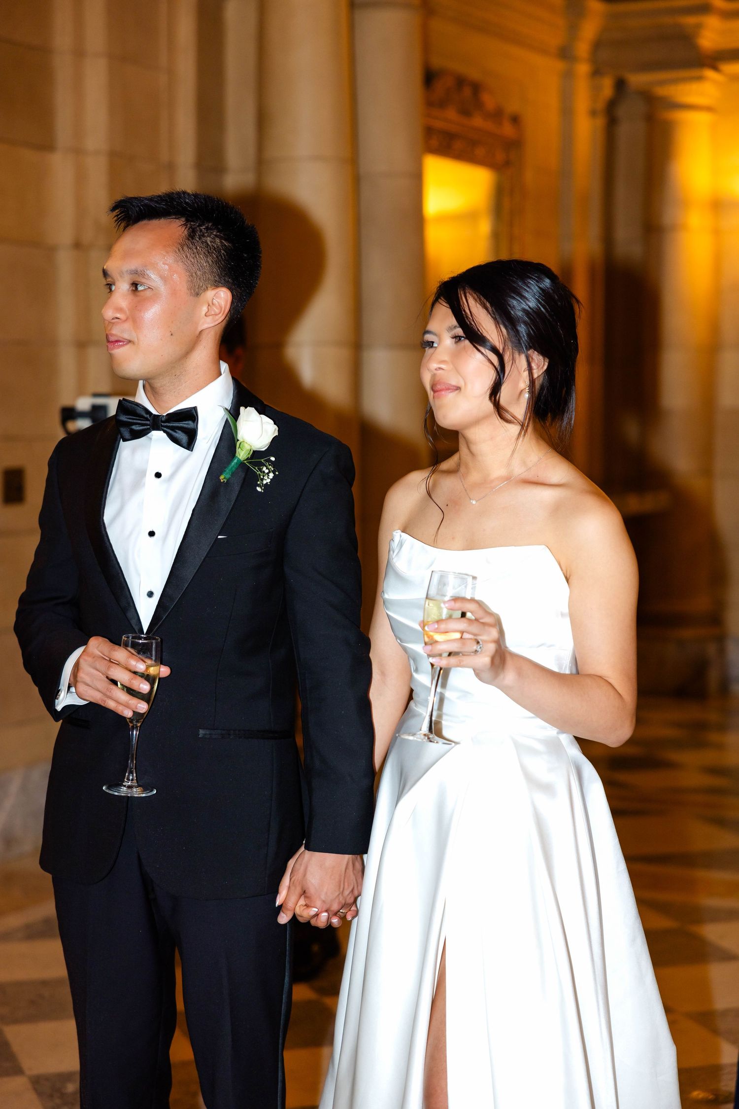 Asian bride and groom in formal wedding attire holding champagne glasses and hands in elegant venue.
