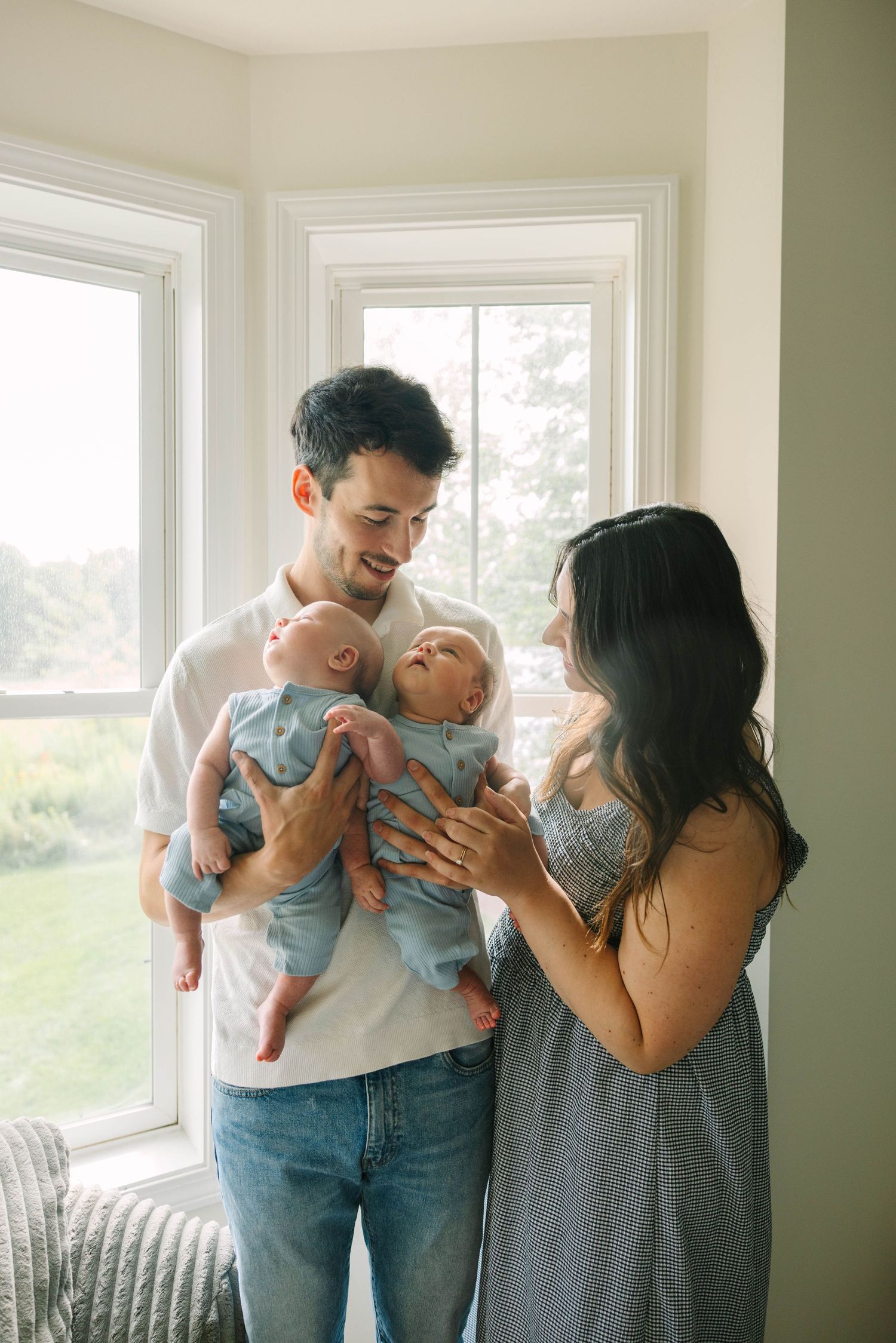 Loving parents admiring their twin babies in blue outfits while standing near sunlit windows.