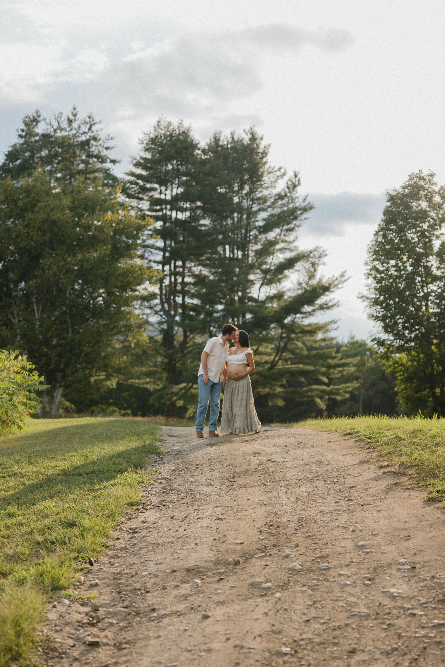 A couple walks hand-in-hand on a dirt path surrounded by lush green trees during an outdoor maternity photoshoot.