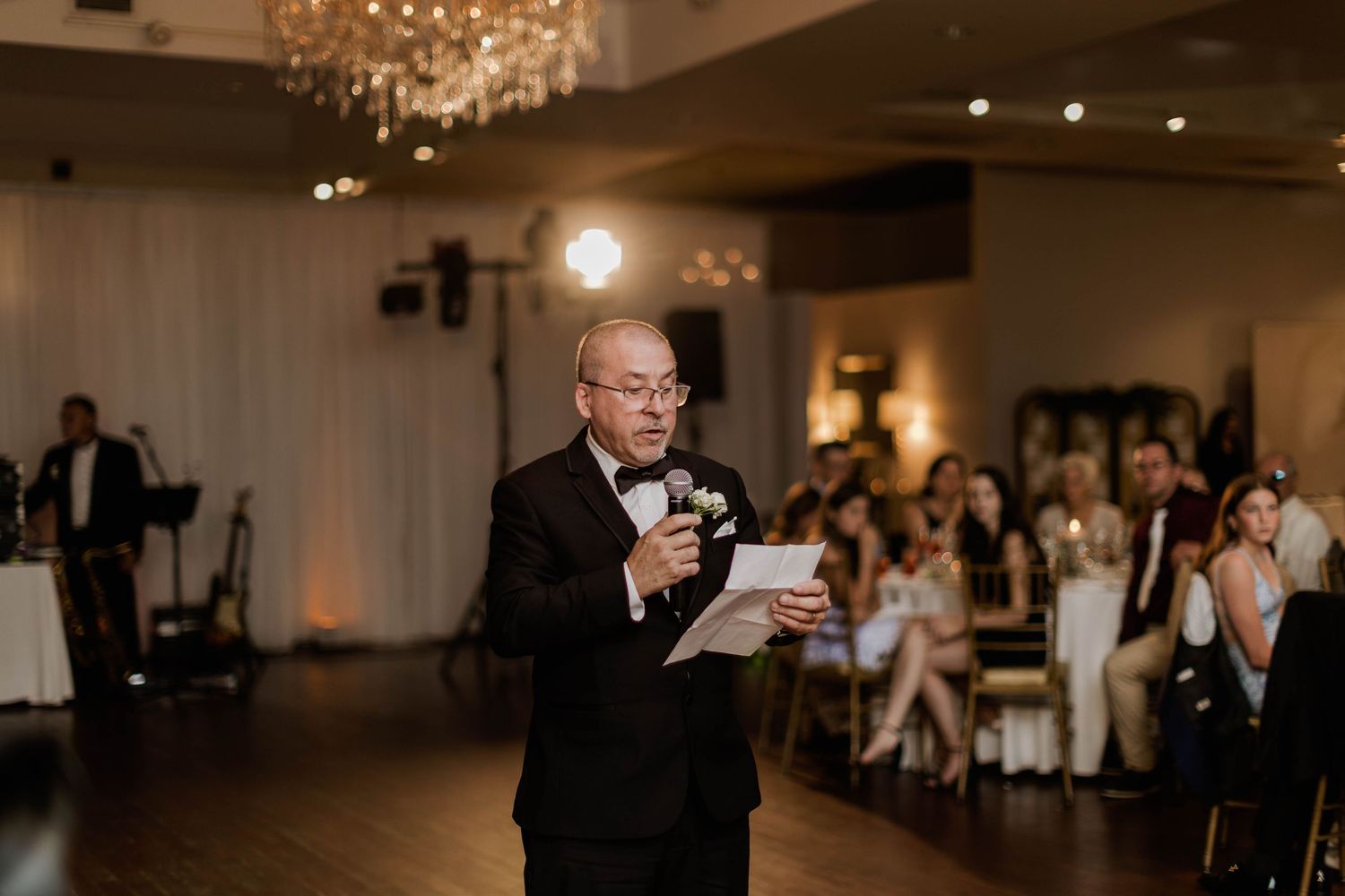 Elderly man in tuxedo delivers a heartfelt speech at a wedding reception in an elegant ballroom.