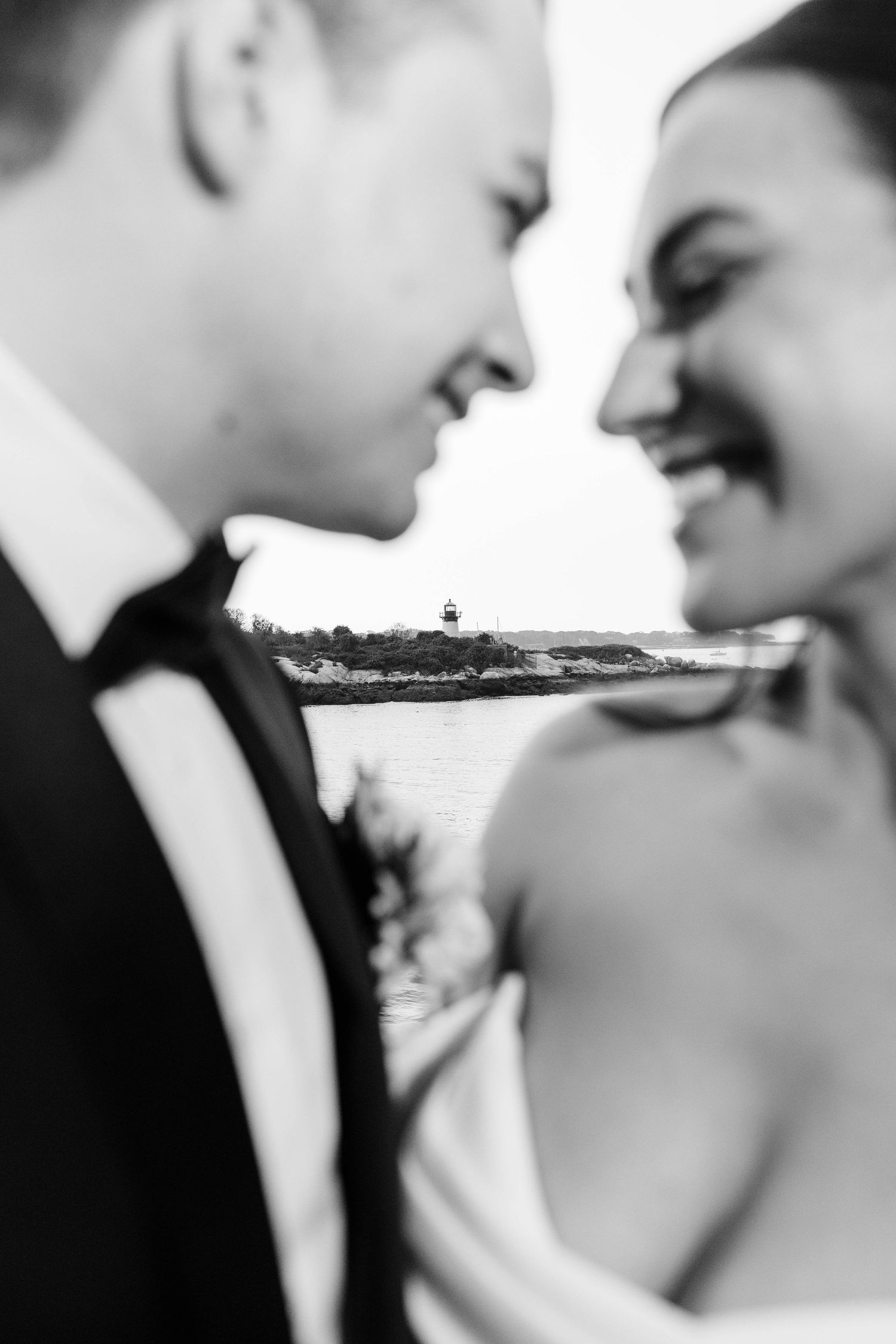 Black and white portrait of smiling bride and groom facing each other with waterfront lighthouse in background.