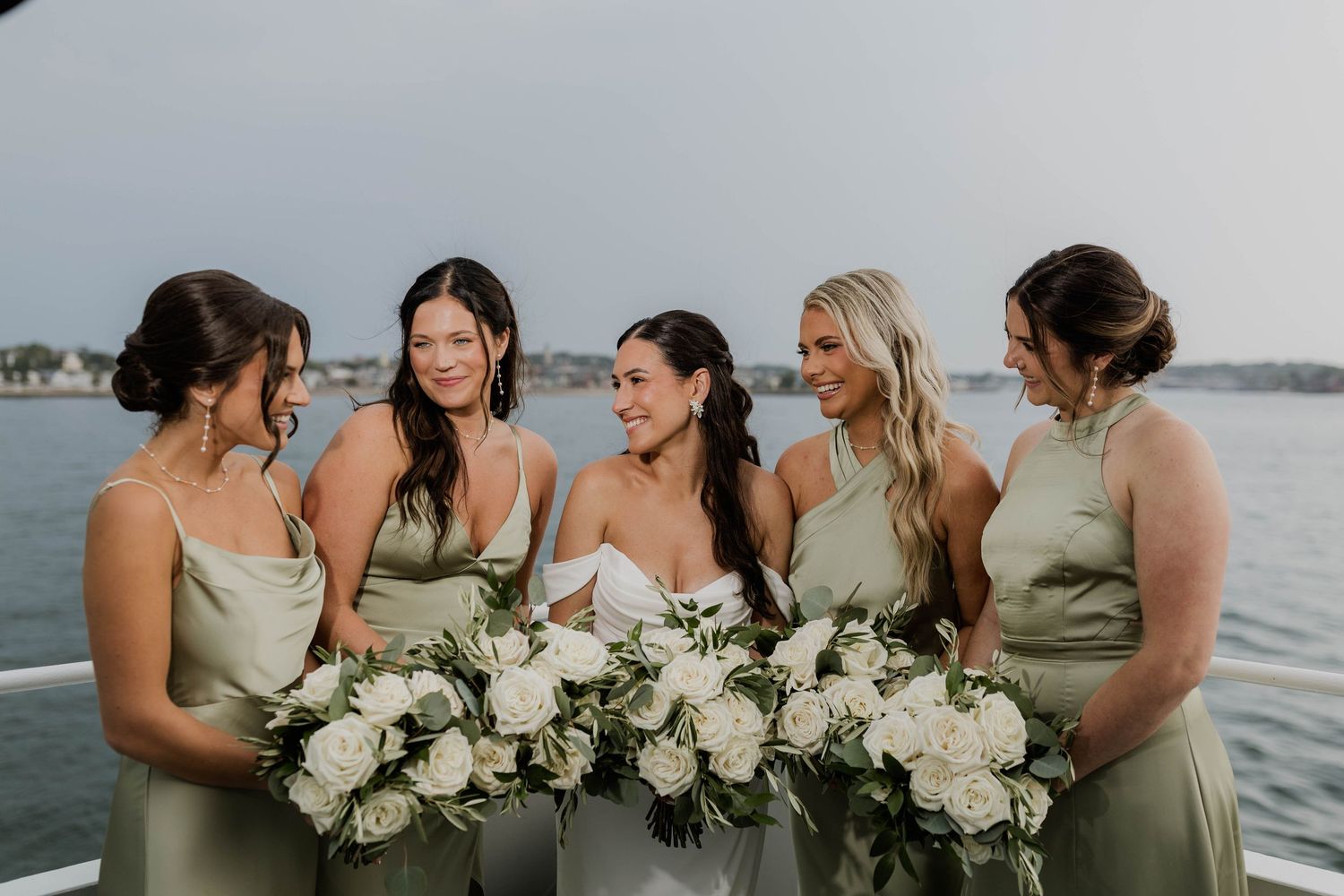 Bride surrounded by four bridesmaids in sage green dresses laughing with white rose bouquets outdoors.