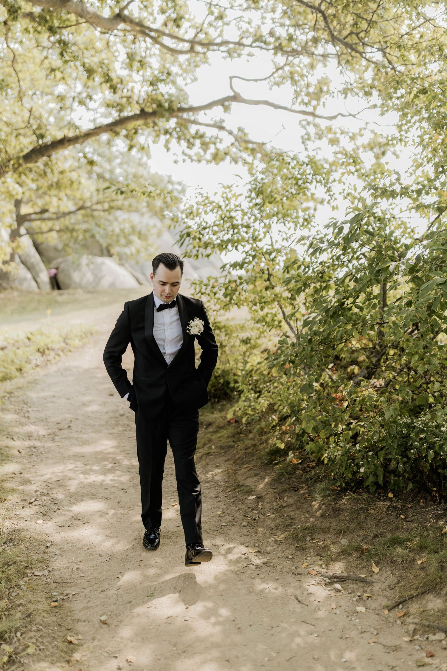 Groom in black tuxedo with bow tie and boutonniere walking along a tree-lined garden path.
