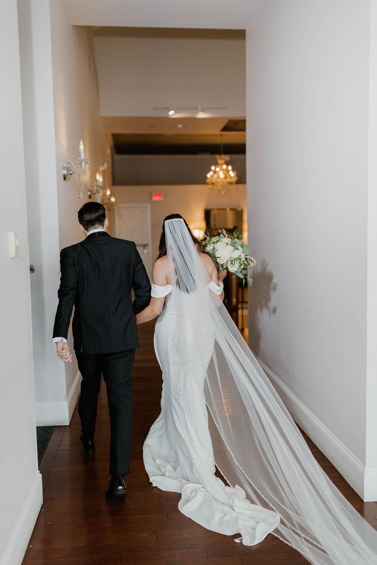 Bride and groom walking hand in hand down hallway, wedding veil trailing behind elegantly.