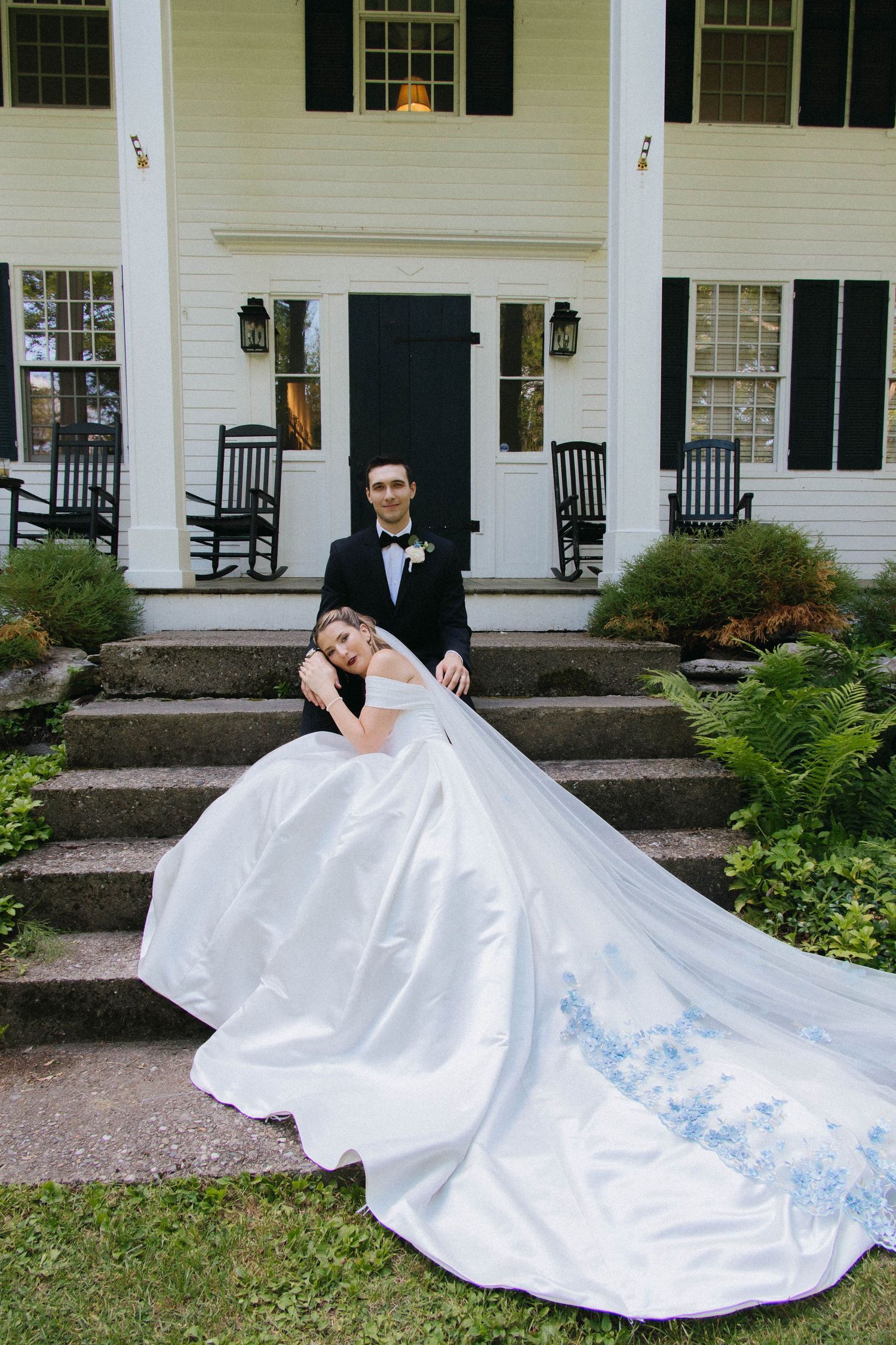 Bride in white ball gown with blue floral details poses with groom in tuxedo on steps of white colonial home.